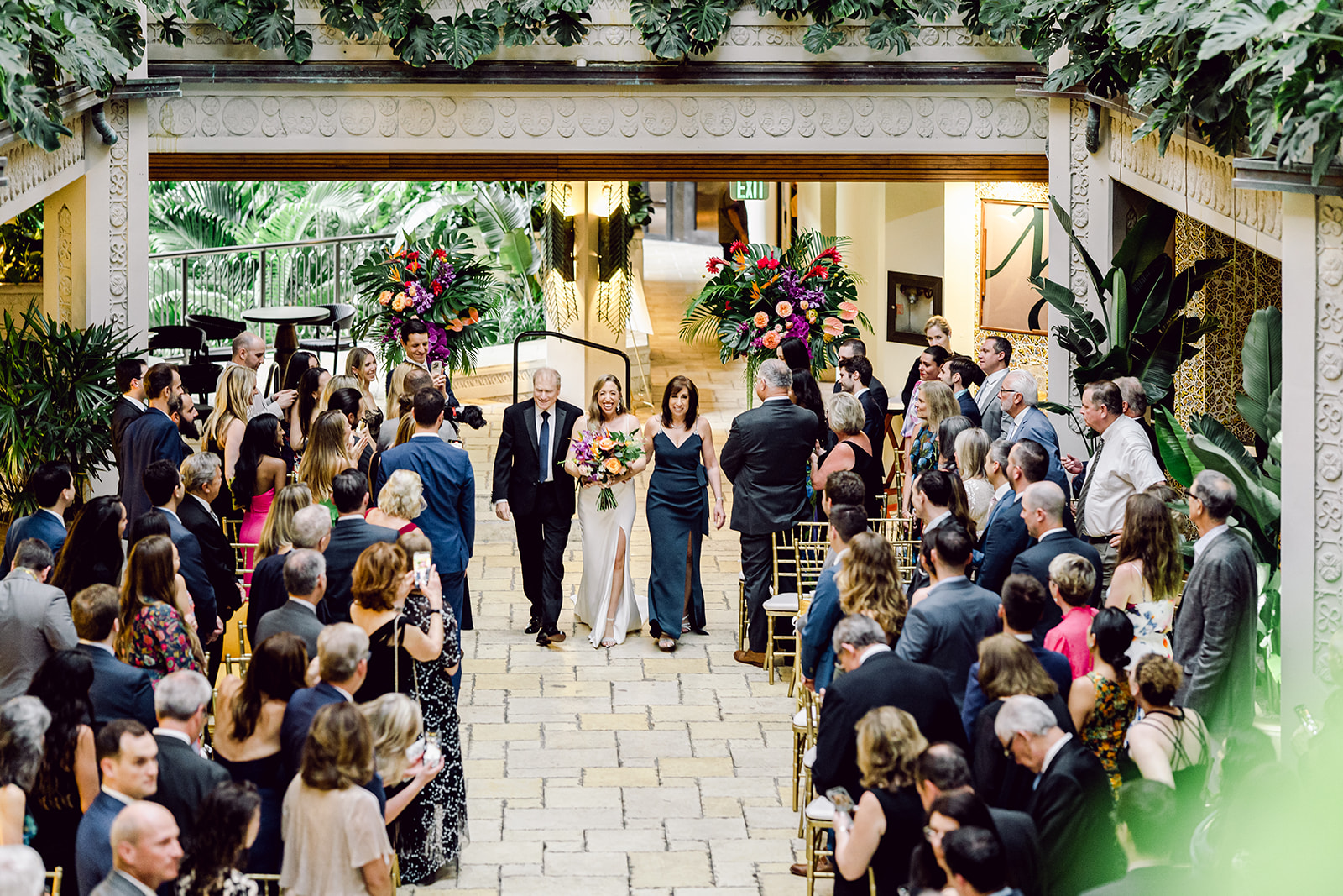 Wide angle of bride with parents walking down aisle at ceremony at Mayfair House Hotel & Garden in Miami on wedding day.