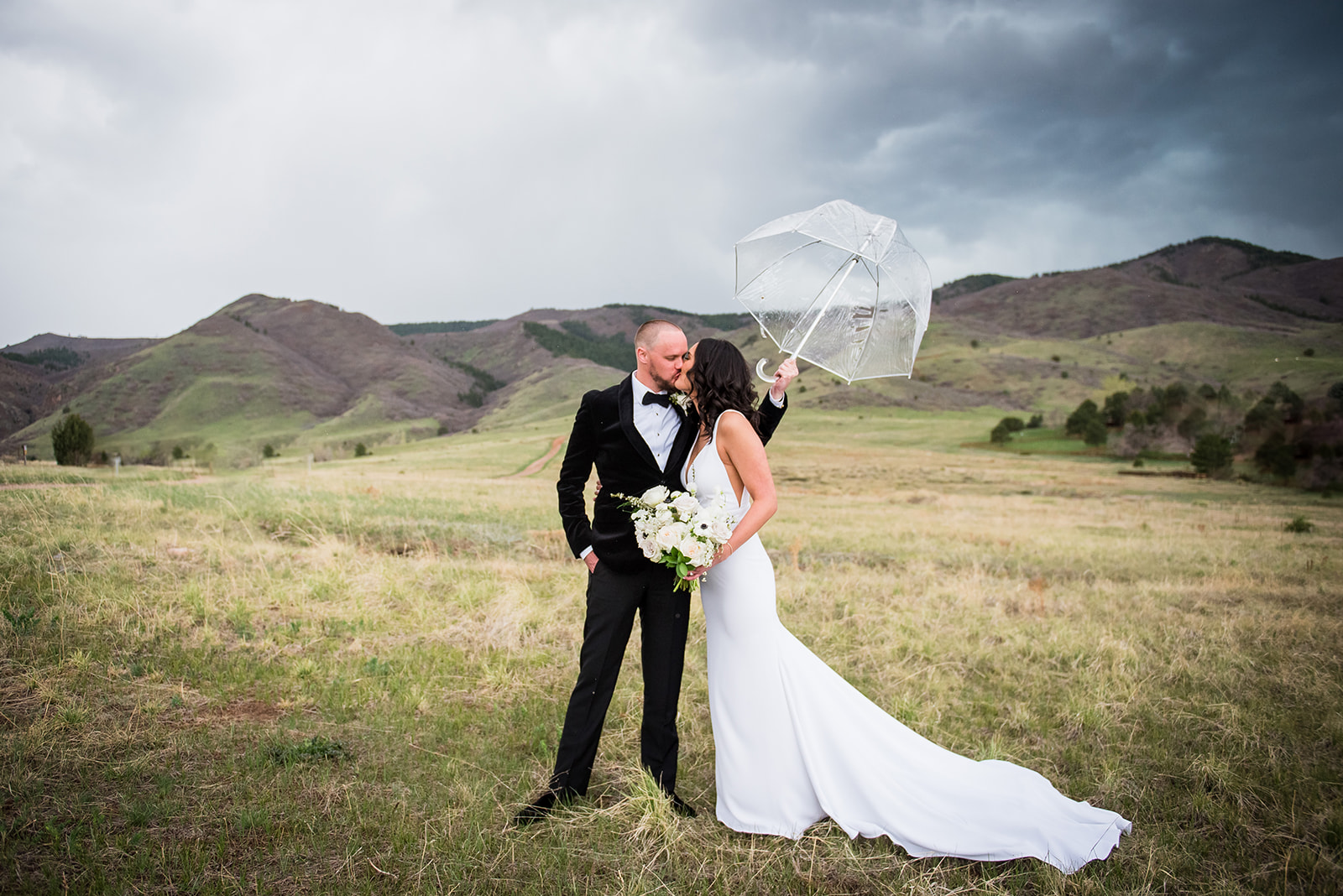 Bride and groom share a kiss in front of the rolling hills of Colorado while groom holds umbrella over them.