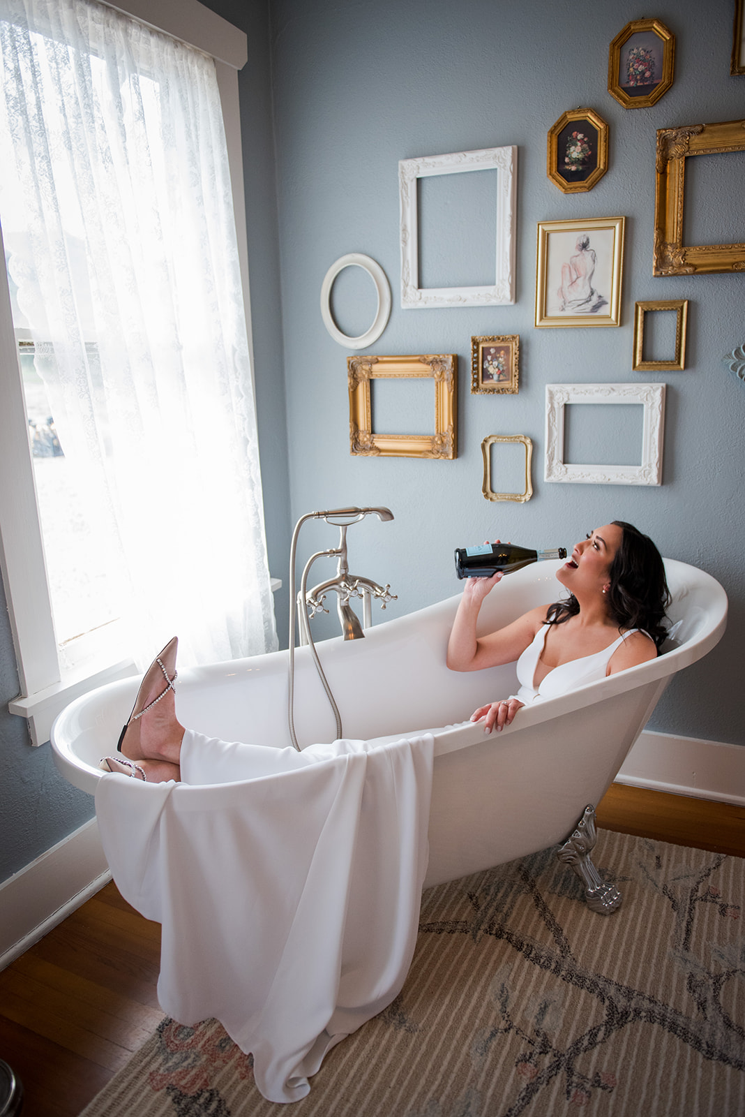 Bride lays in clawfoot bathtub in her wedding dress drinking a bottle of champagne.