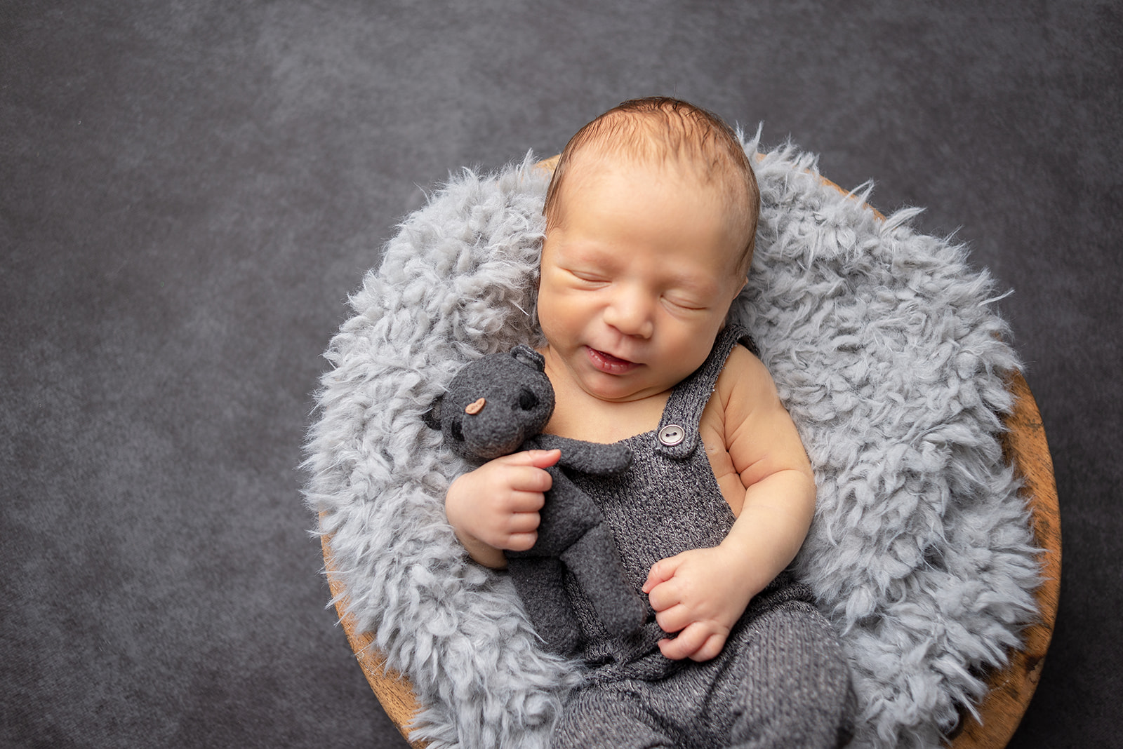 A baby boy sleeping on a gray fuzzy blanket placed inside a wooden bowl in Celesta Champagne Photography studio