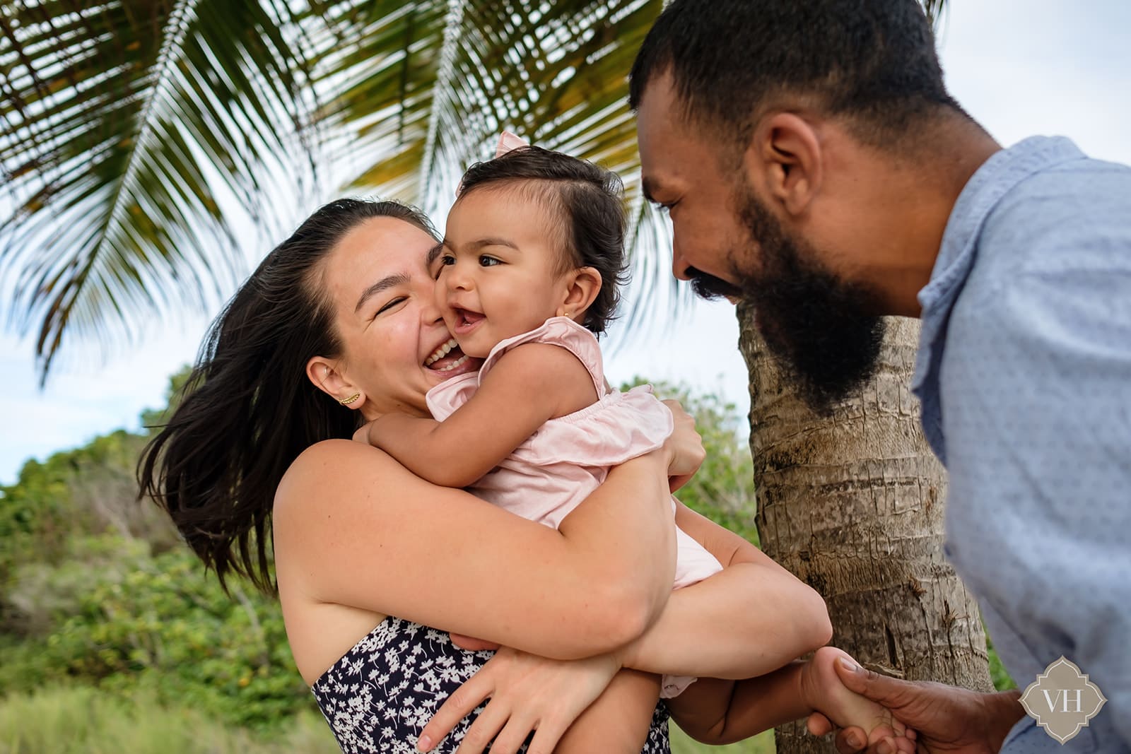 mother and father laughing and cuddling baby girl under a coconut tree at jabbawock beach antigua 