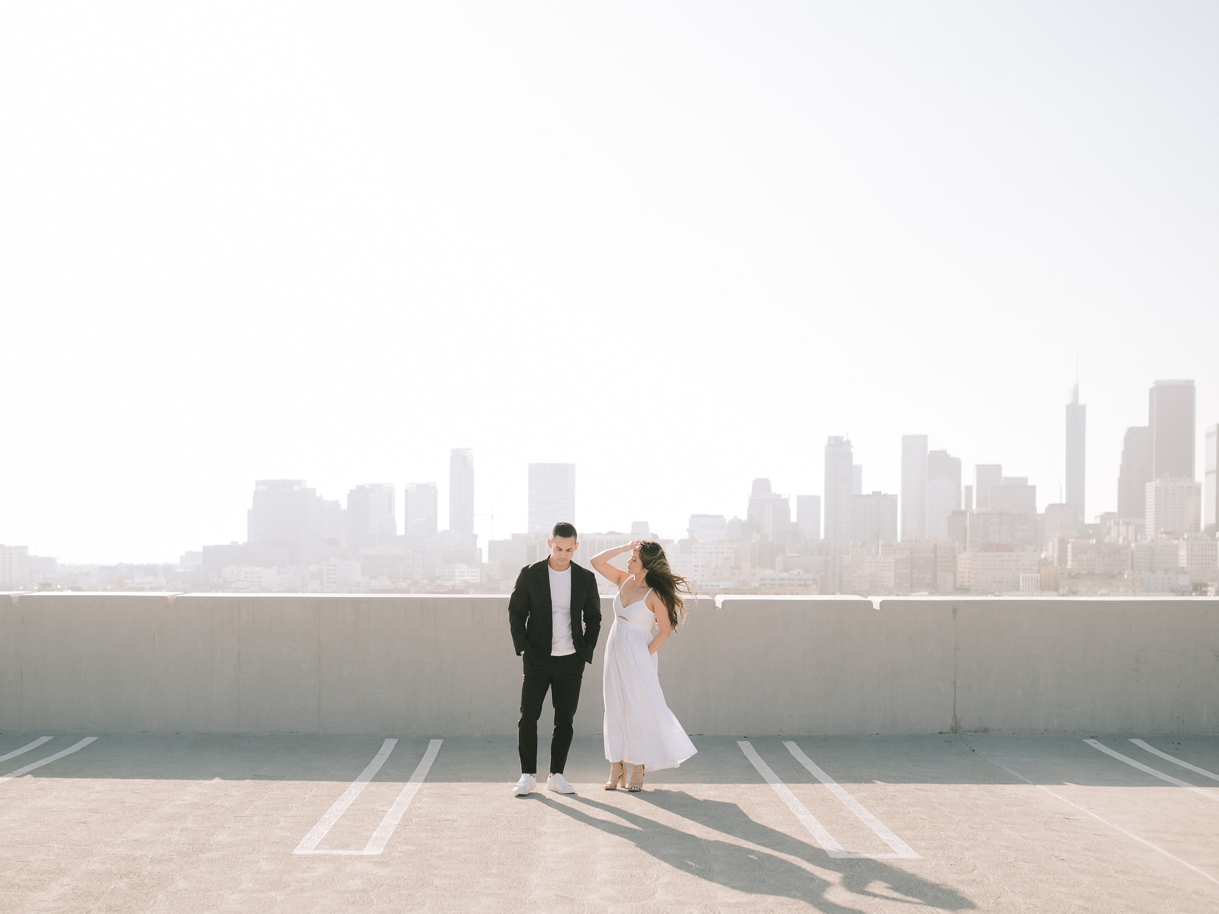 An engaged couple stands stylishly in front of the downtown Los Angeles skyline. 