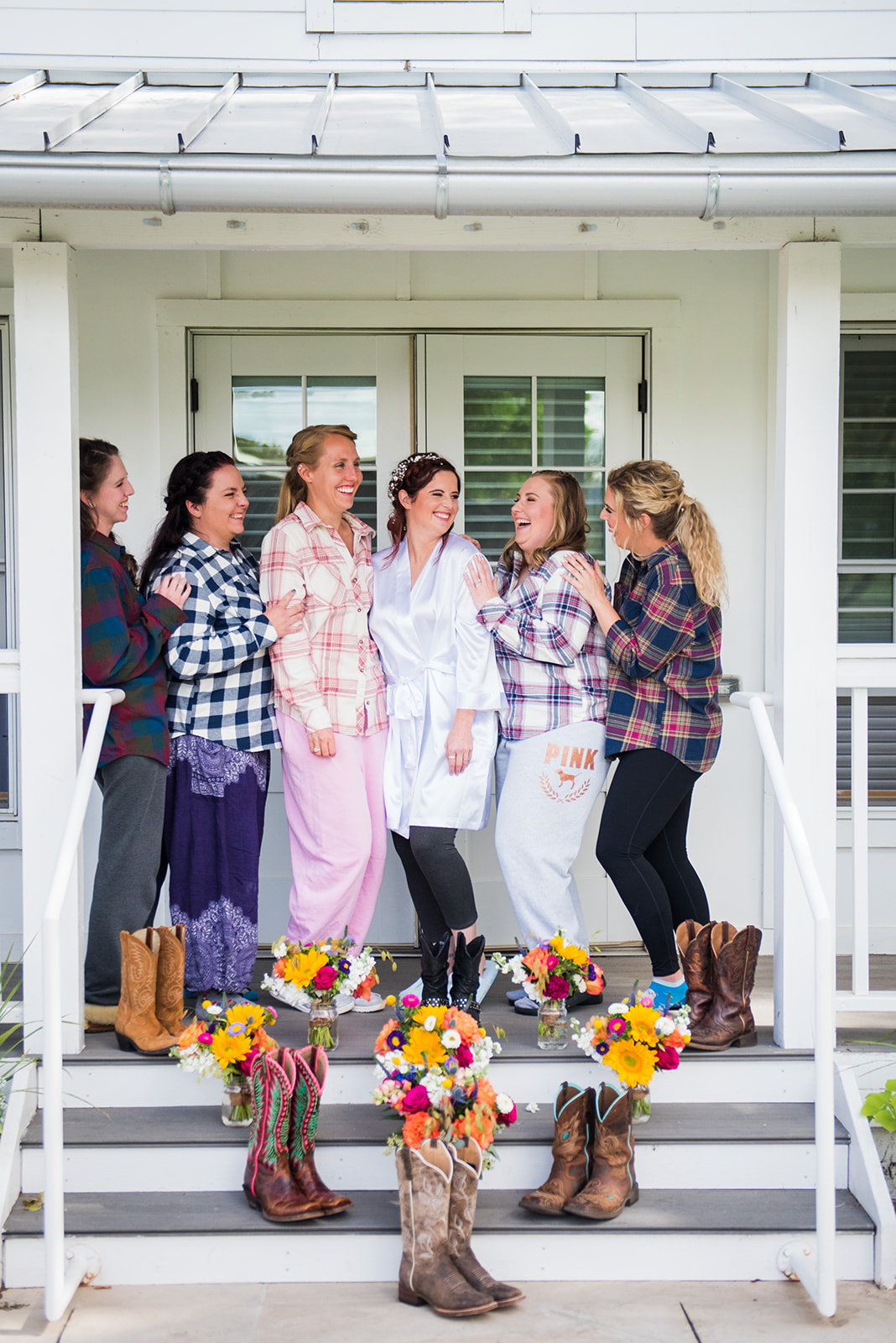 Bride and bridesmaids stand on front porch smiling at each other. Bridesmaids are wearing different color flannel shirts