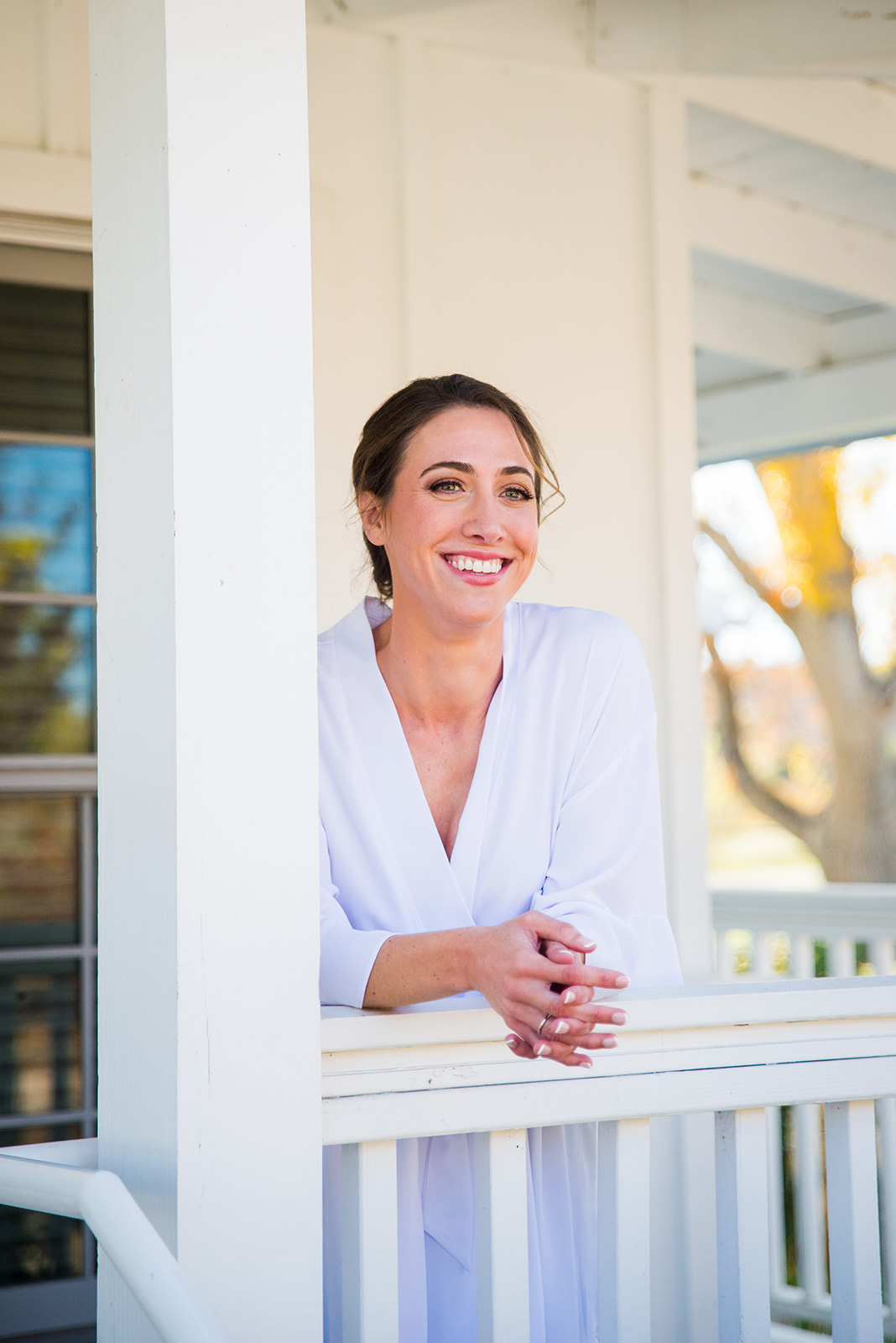 Bride stands on front porch, leaning over the railing, smiling and wearing a white silk robe.