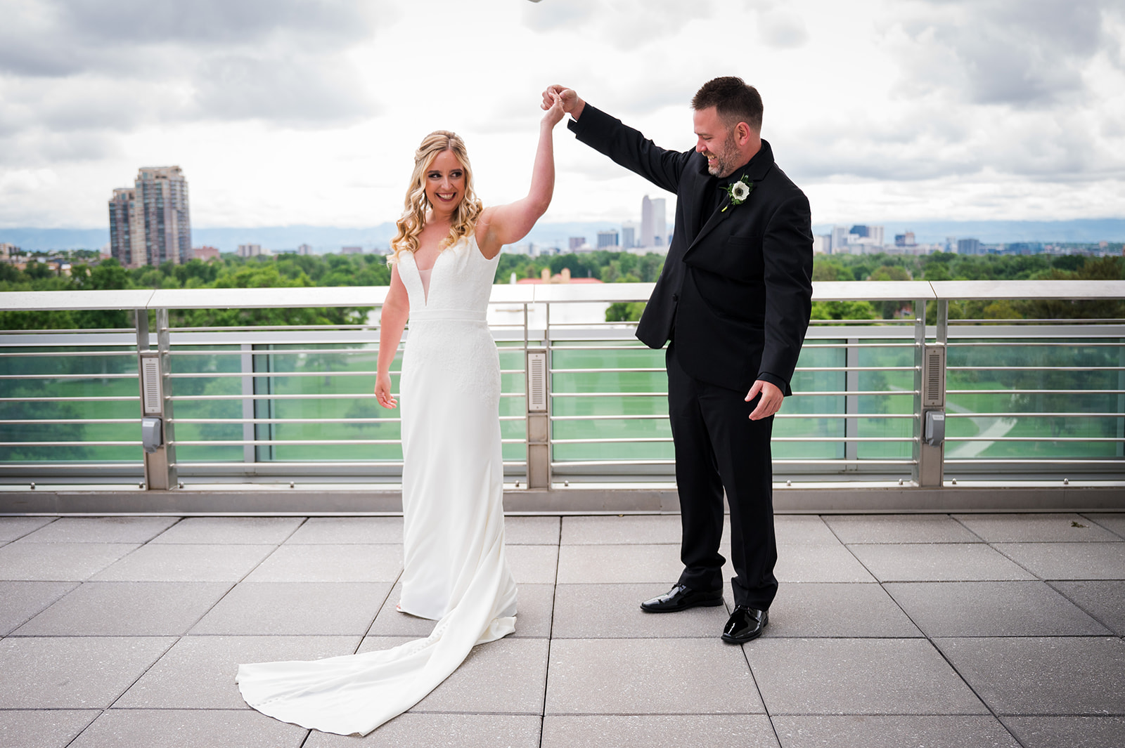 Groom spins bride around on a rooftop with Denver city skyline in the background.