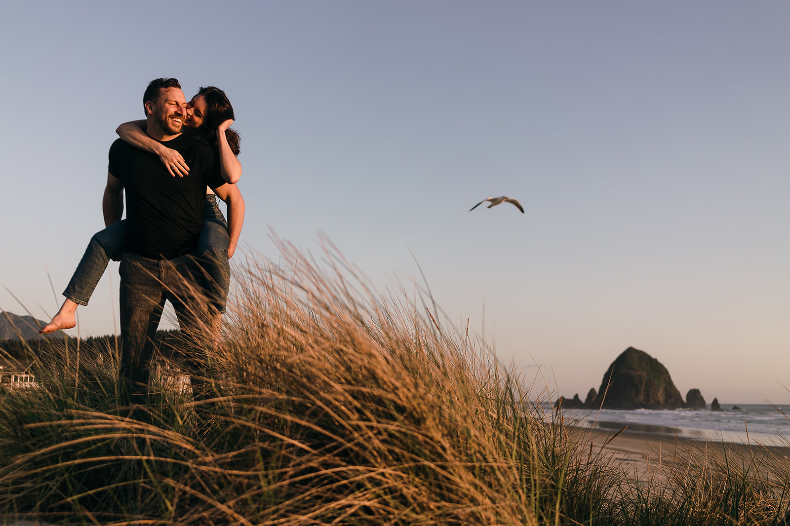 couple climbs over beach grass on sand dunes during Cannon Beach Engagement session