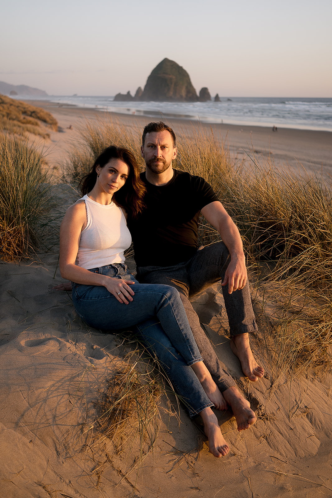 couple poses for engagement photos at Cannon Beach with Haystack Rock behind them