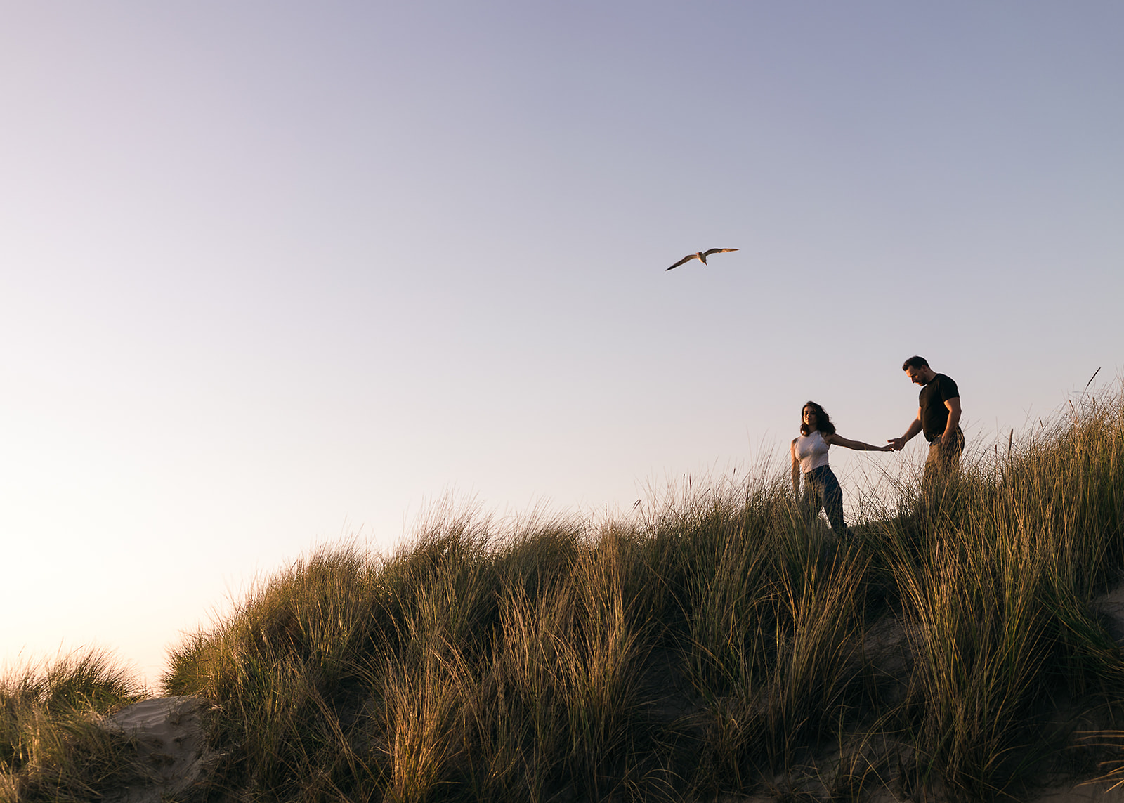couple walks along cannon beach dunes between sea grass during engagement session