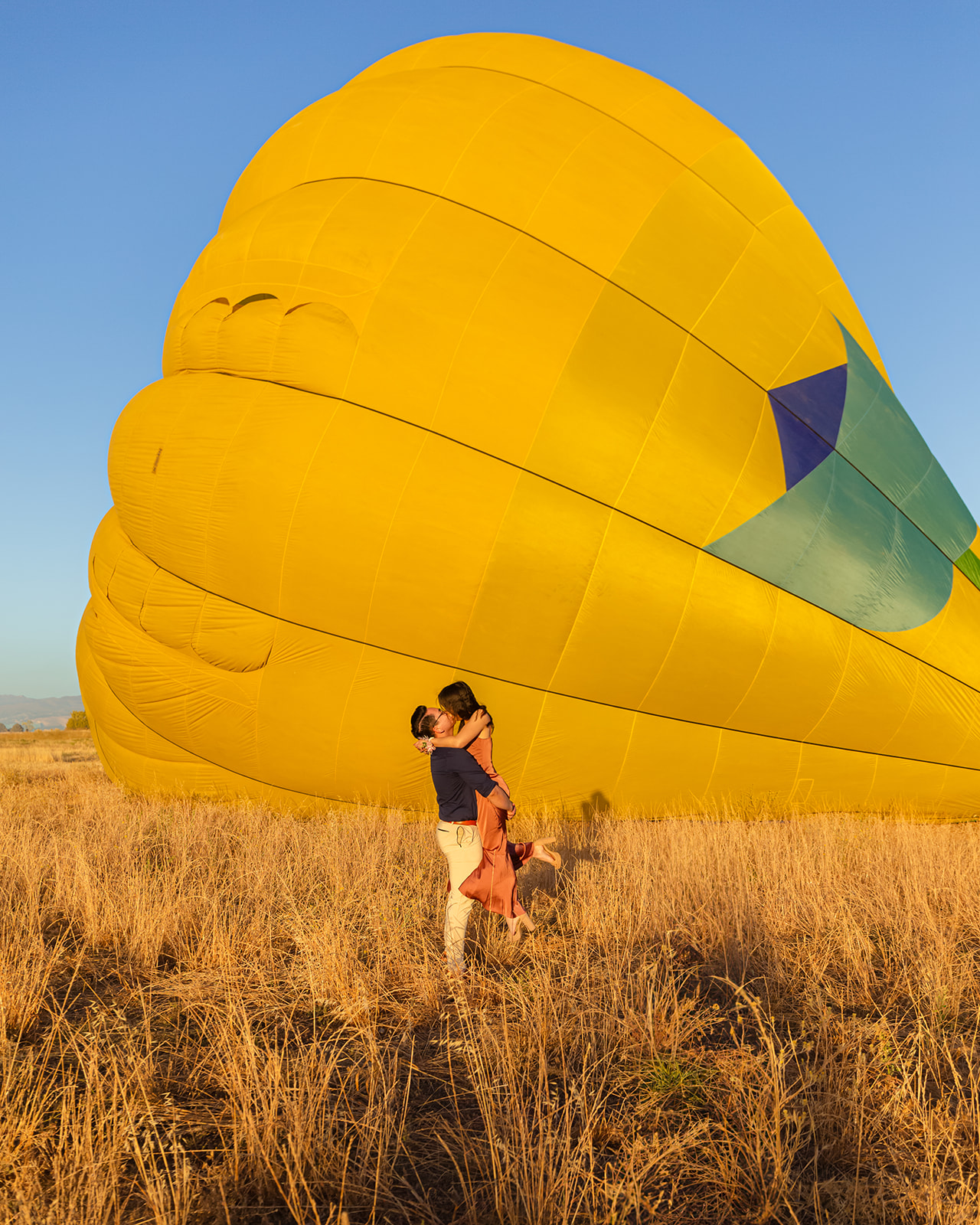 nonbinary person lifting their fiancée in front of a yellow hot air balloon