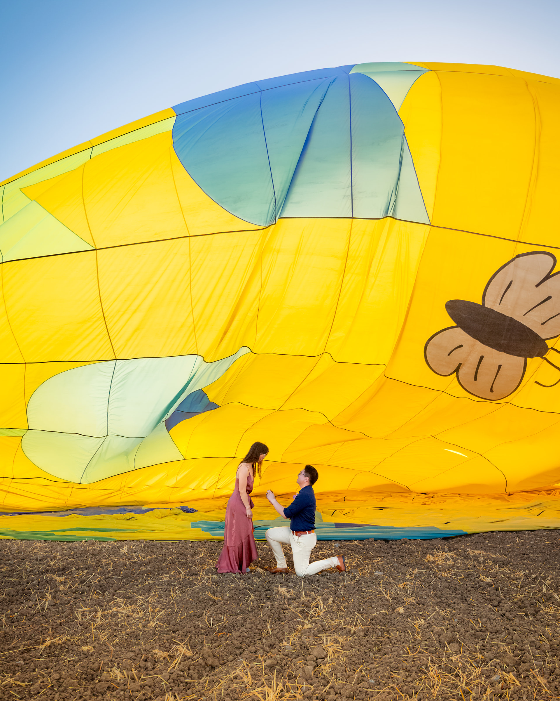 nonbinary person proposing to their fiancée in front of a yellow hot air balloon