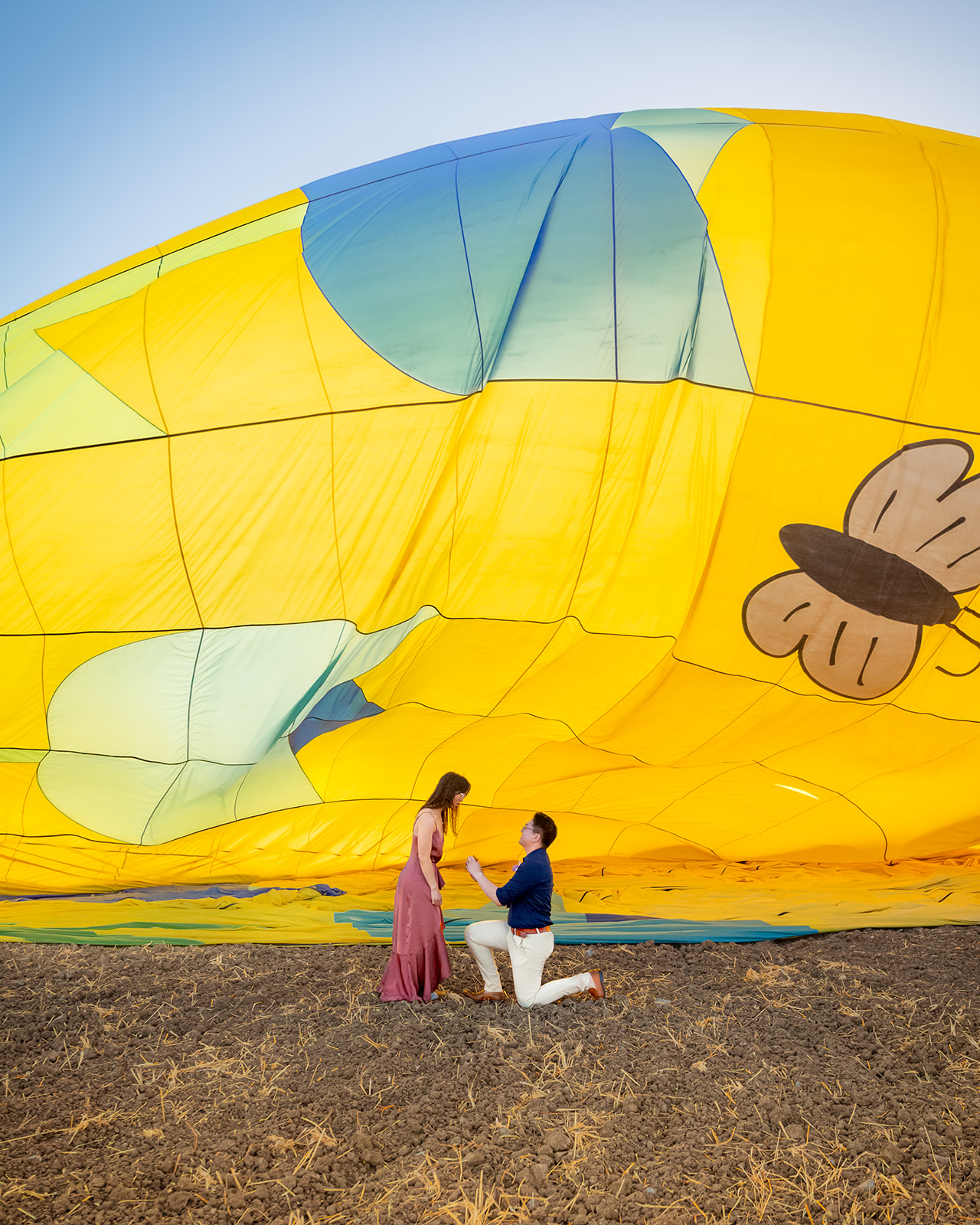 nonbinary person proposing to their girlfriend in front of yellow hot air balloon