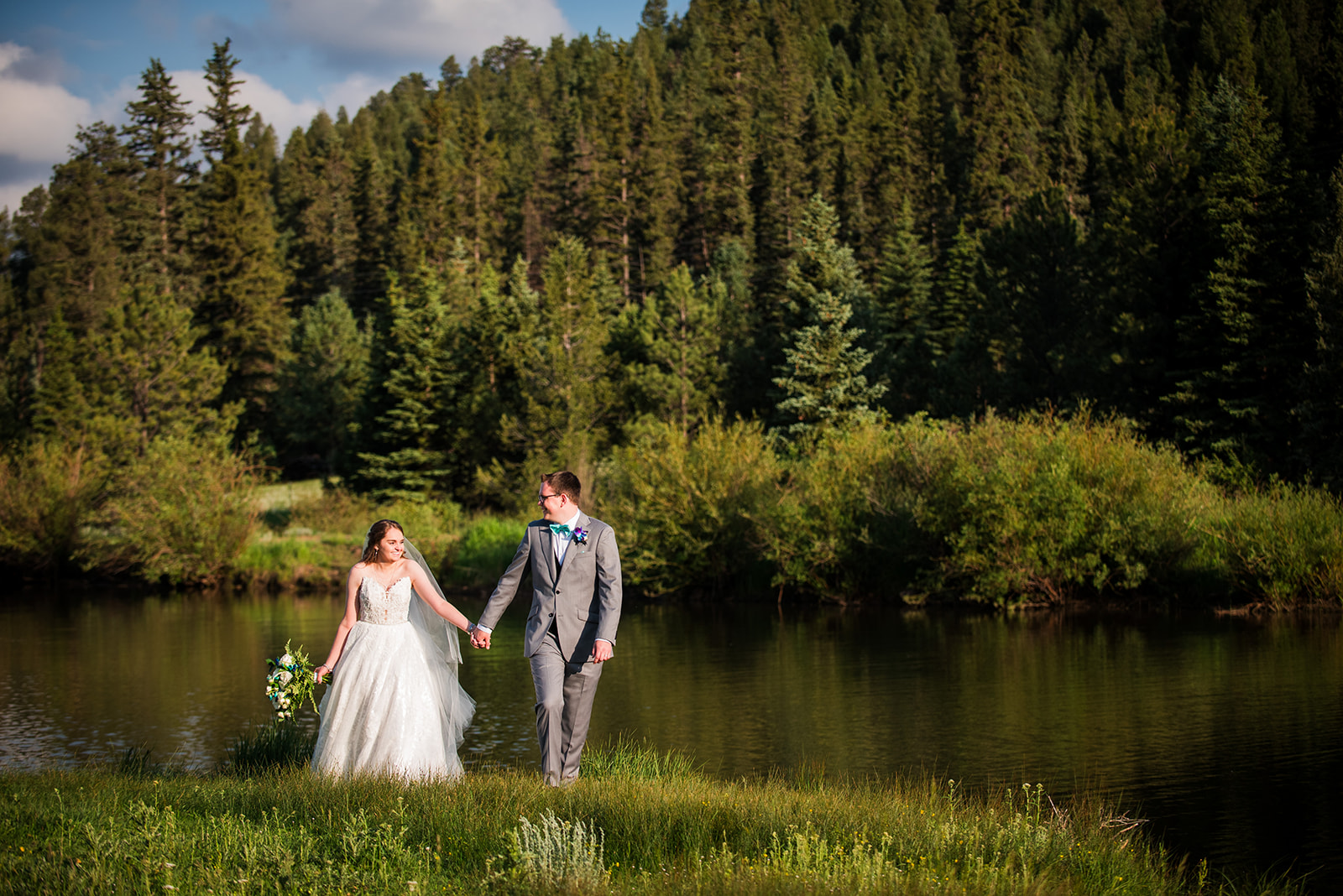 The bride and groom walk toward the camera hand-in-hand with a lake and pine trees in the background.
