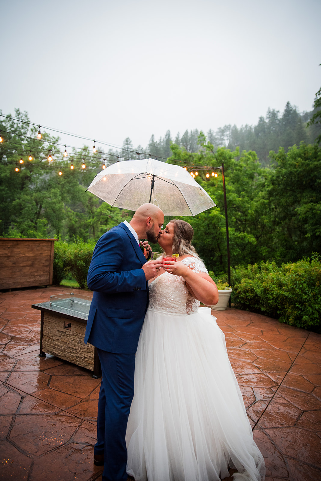 The bride and groom share a kiss in the rain under a clear umbrella.