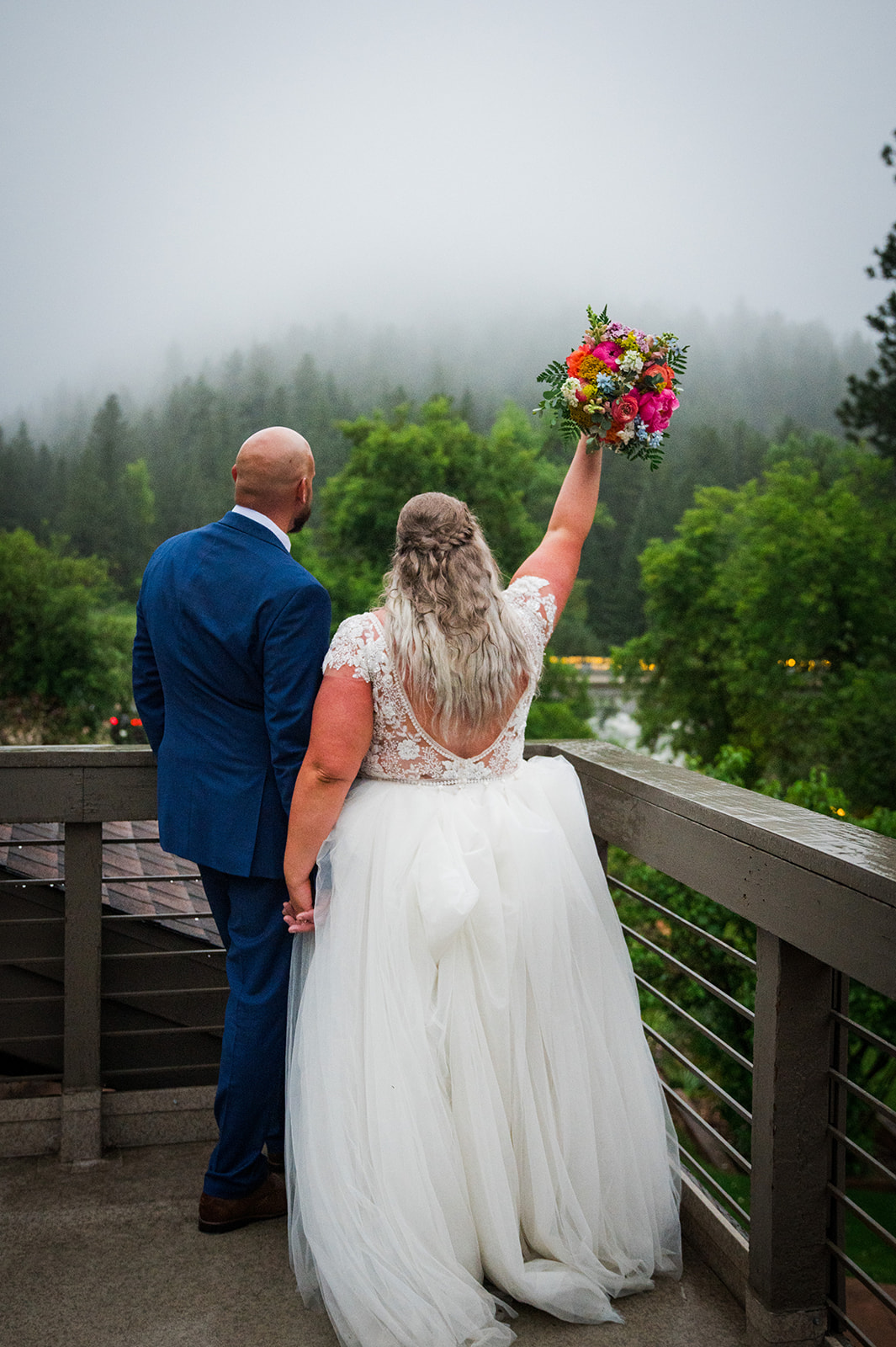The bride and groom stand on a terrace overlooking a foggy view.