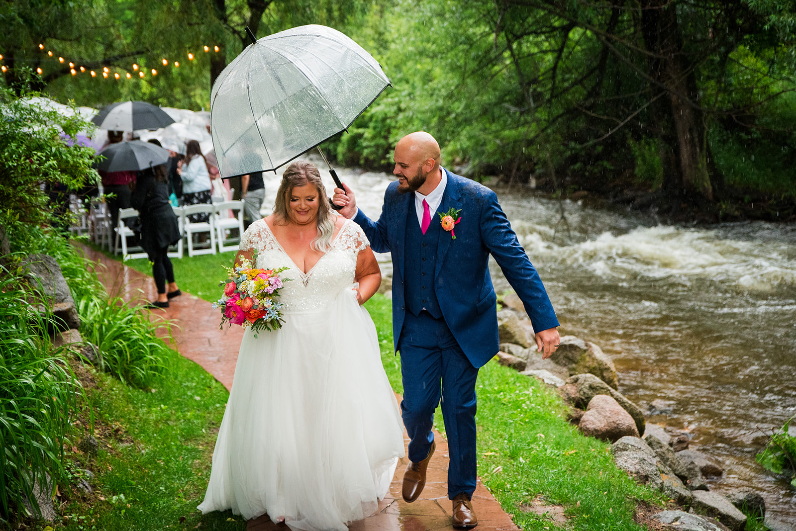 The bride and groom walk toward the camera hand-in-hand as the groom holds a clear umbrella above them.