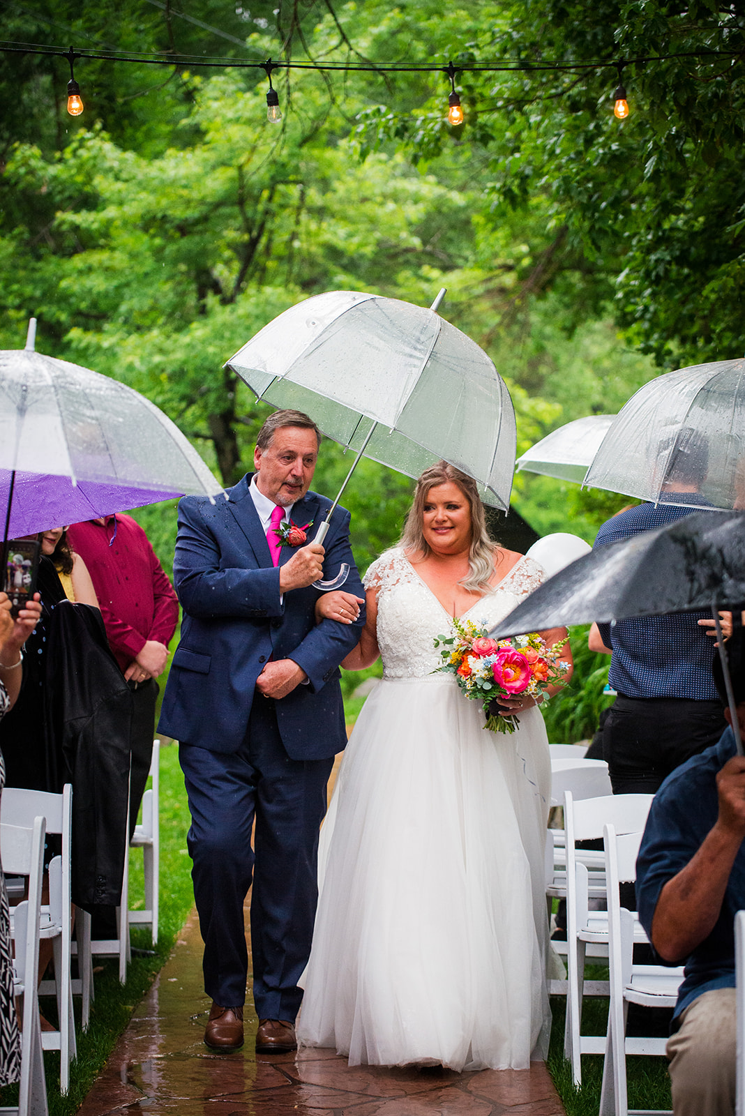 The bride's father walks her up the aisle in the rain, holding a clear umbrella.