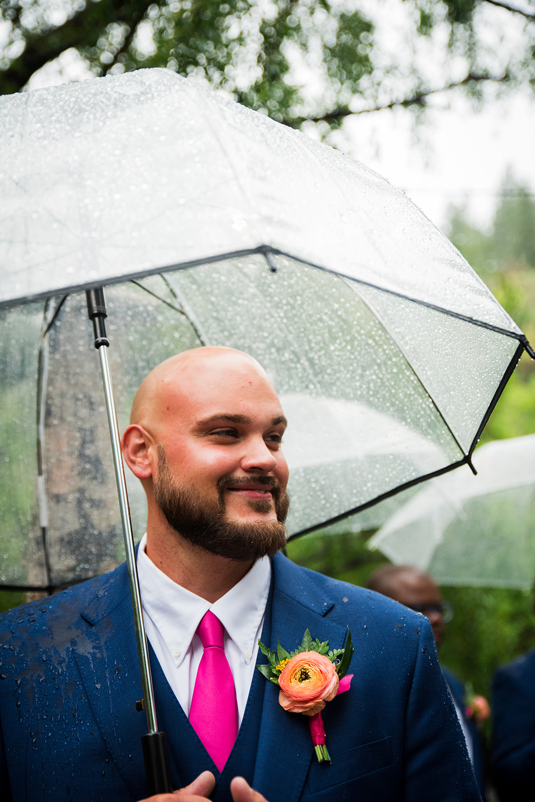 The groom stands looking at his bride walking down the aisle, holding a clear umbrella.