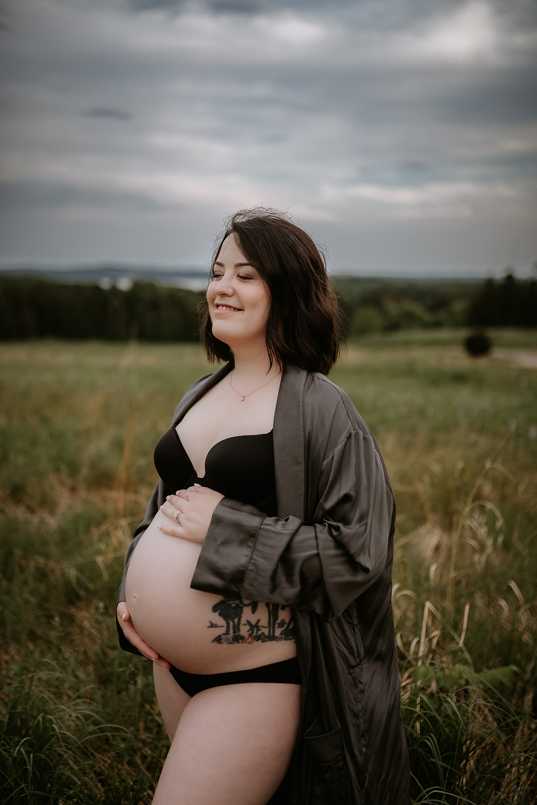Pregnant mother in a dark gray satin robe holding her belly atop a hill of grasses and wildflowers