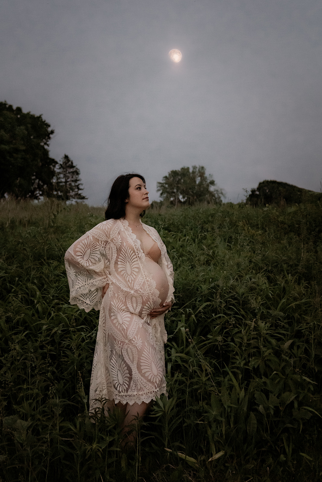 Pregnant mother in a long lace robe touching her belly in the woods under the moon
