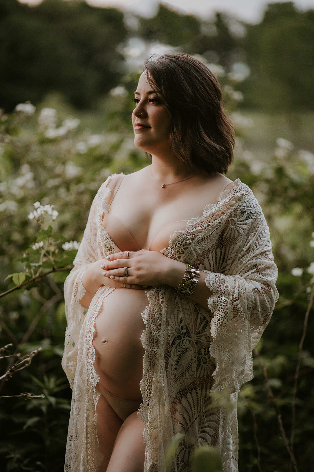 Pregnant woman in an ivory lace robe with her hands placed on her belly next to blackberry bushes