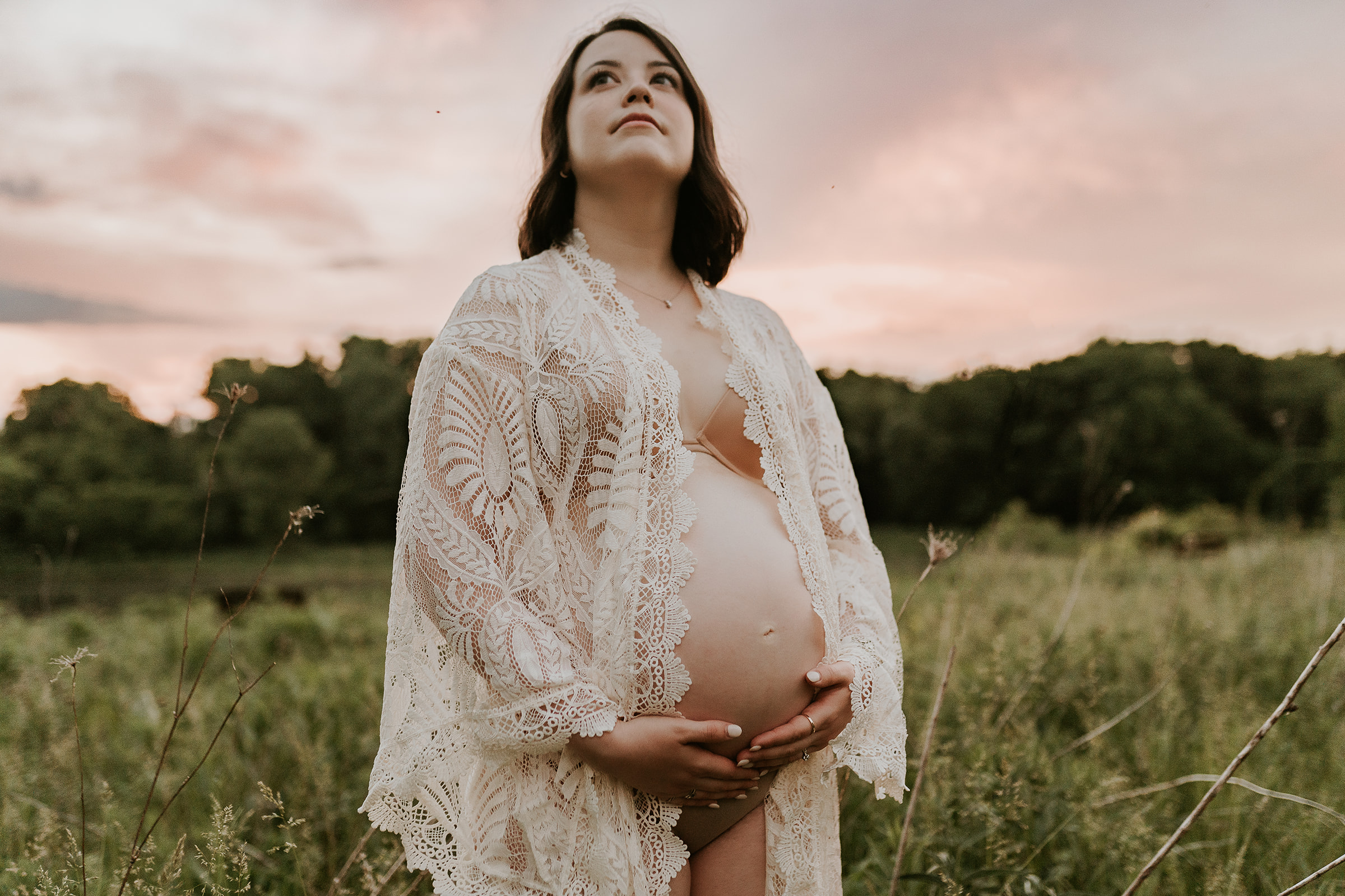 Pregnant woman in an ivory lace robe holding her belly during a cotton candy sunset