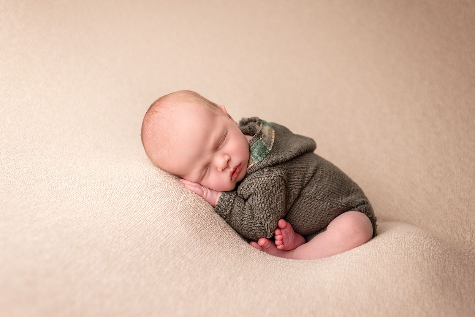 A baby boy in a green knit romper squished together on a beige backdrop sleeping