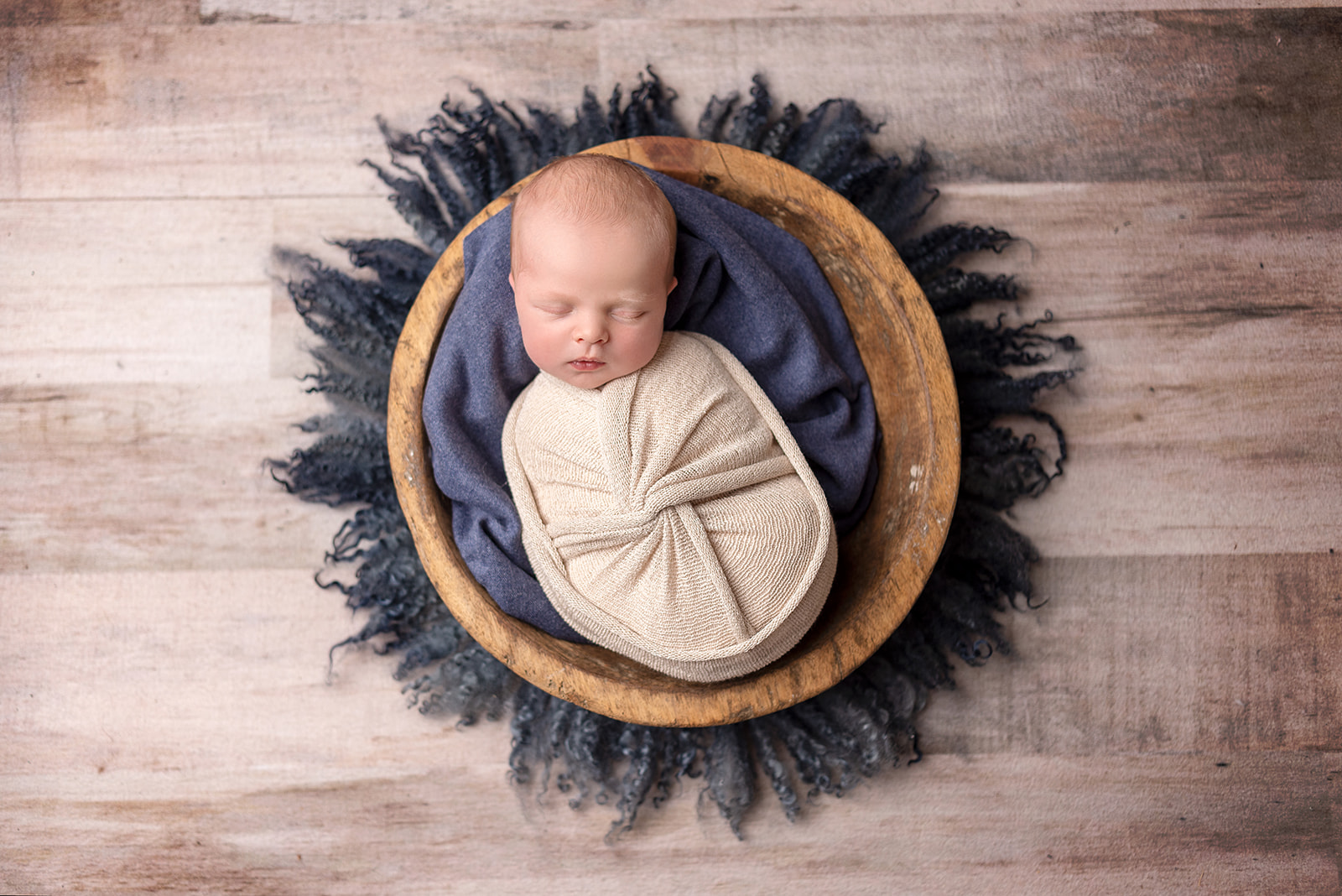 A newborn boy wrapped up in a cream knit swaddle sleeping in a round wooden bowl