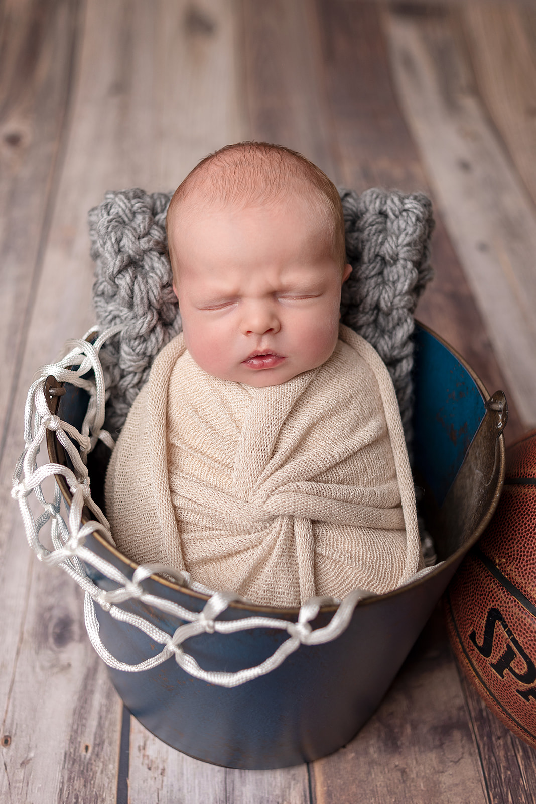 A newborn boy wrapped up in a cream knit swaddle sleeping in a bucket and basketball netting with a basketball nearby