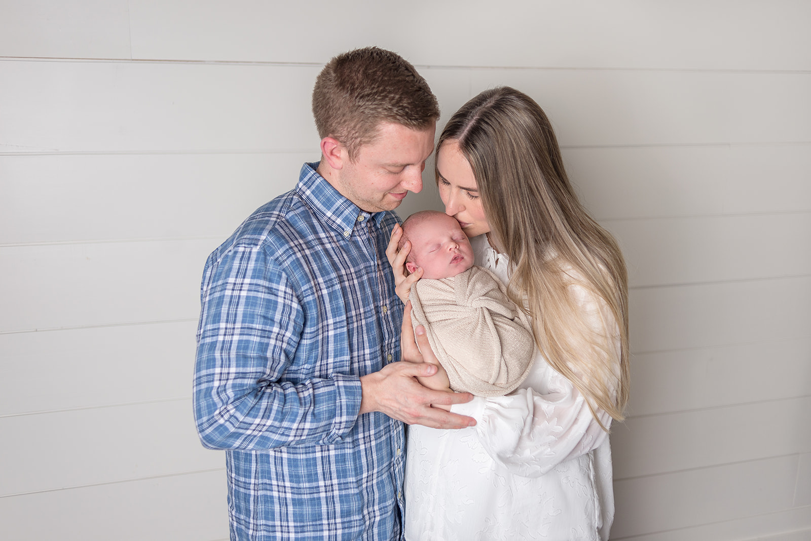A new mom & dad holding her newborn son in a cream swaddle in a all white backdrop studio in Carthage, Missouri