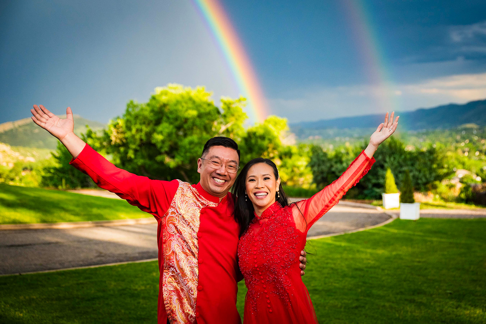 Couple dressed in traditional Vietnamese outfits raises their arms in celebration with a double rainbow behind them.