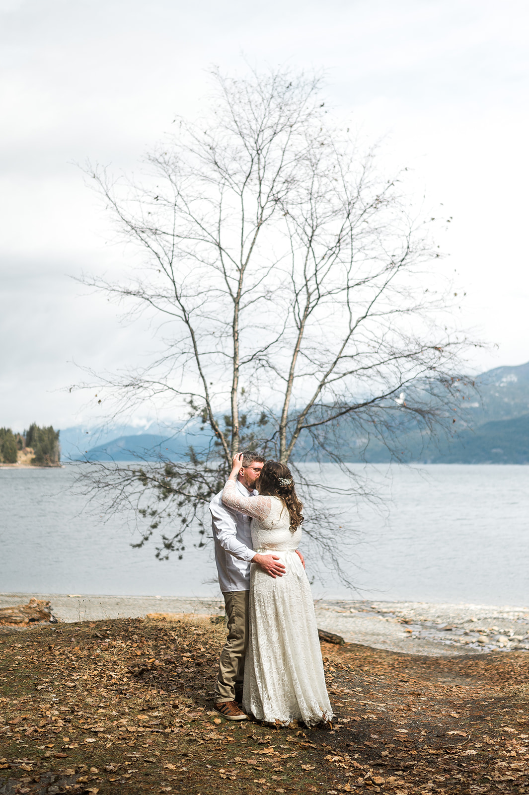The bride and groom sharing a kiss, framed by a tree in front of the lake.