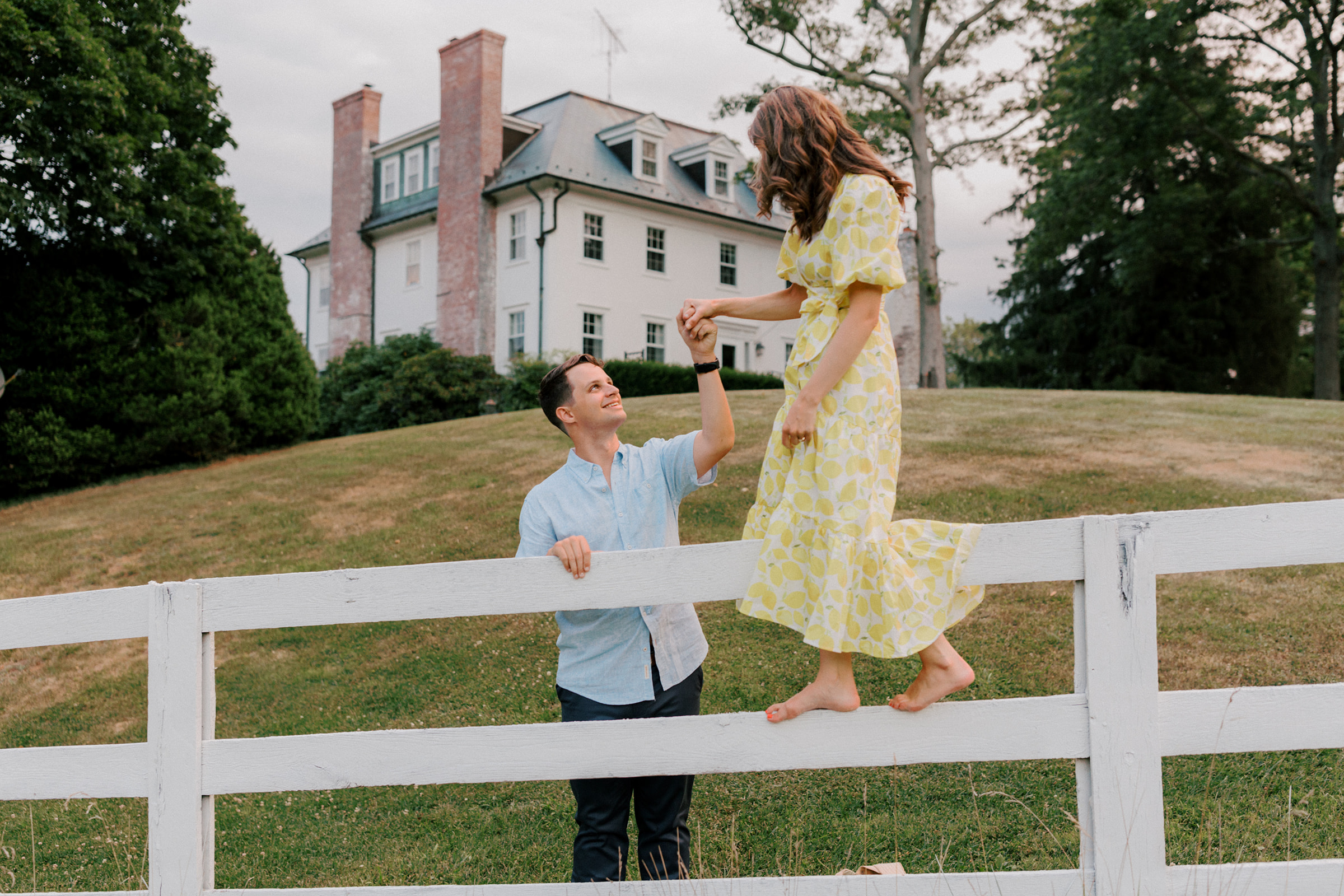 A couple climbs over fence during engagement photos in front of a country farm house estate.