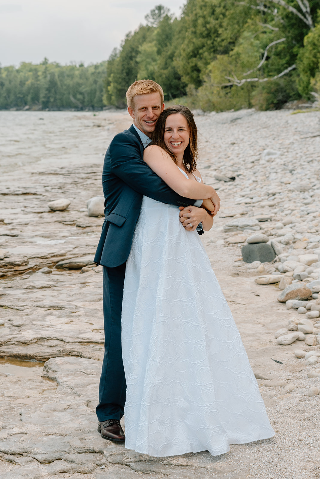 Husband wearing navy blue suite hugs his wife from behind on the shores of Lake Michigan in Door County