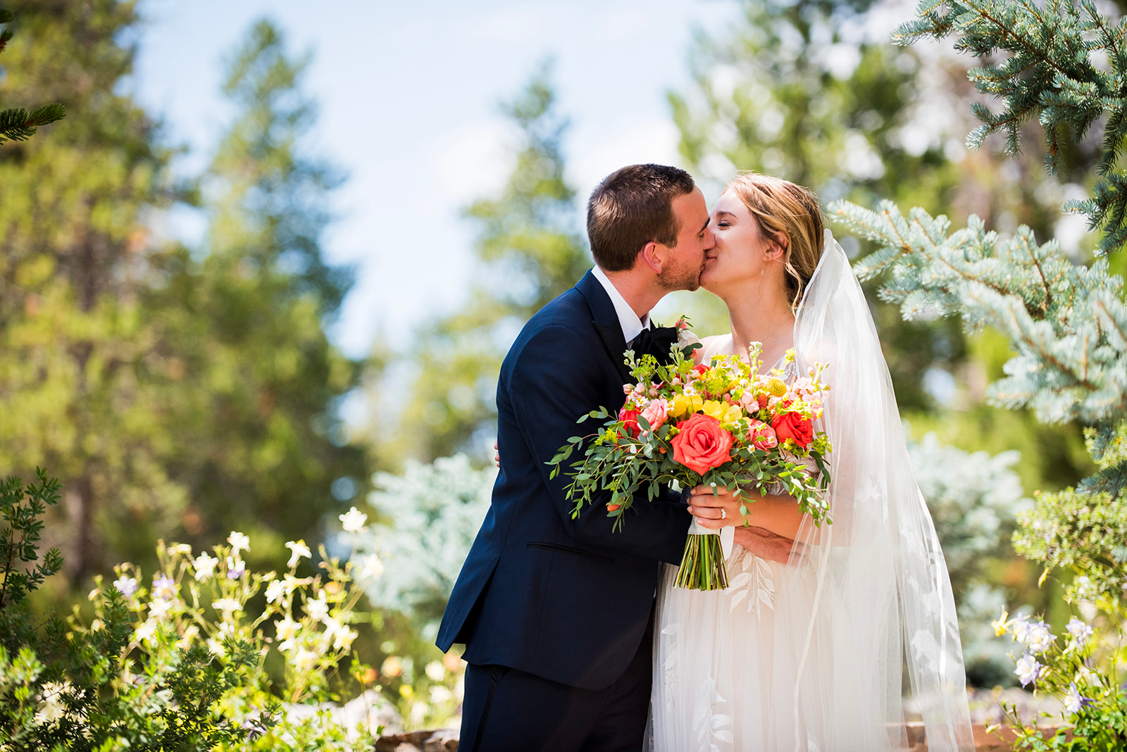 The bride and groom share a kiss as she holds her colorful summer bouquet.