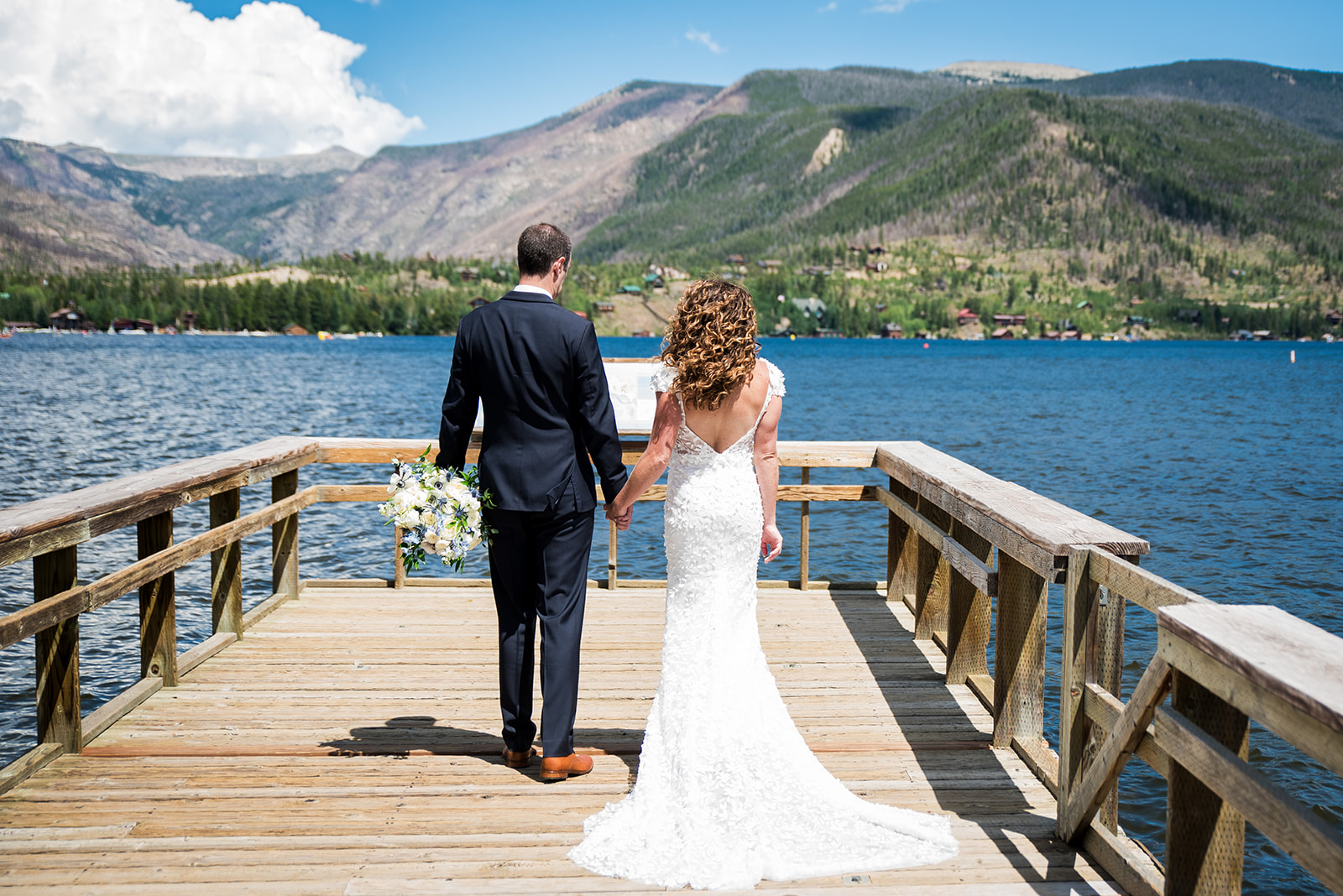 Bride and groom walk hand in hand away from the camera with lake and mountain views in the background.