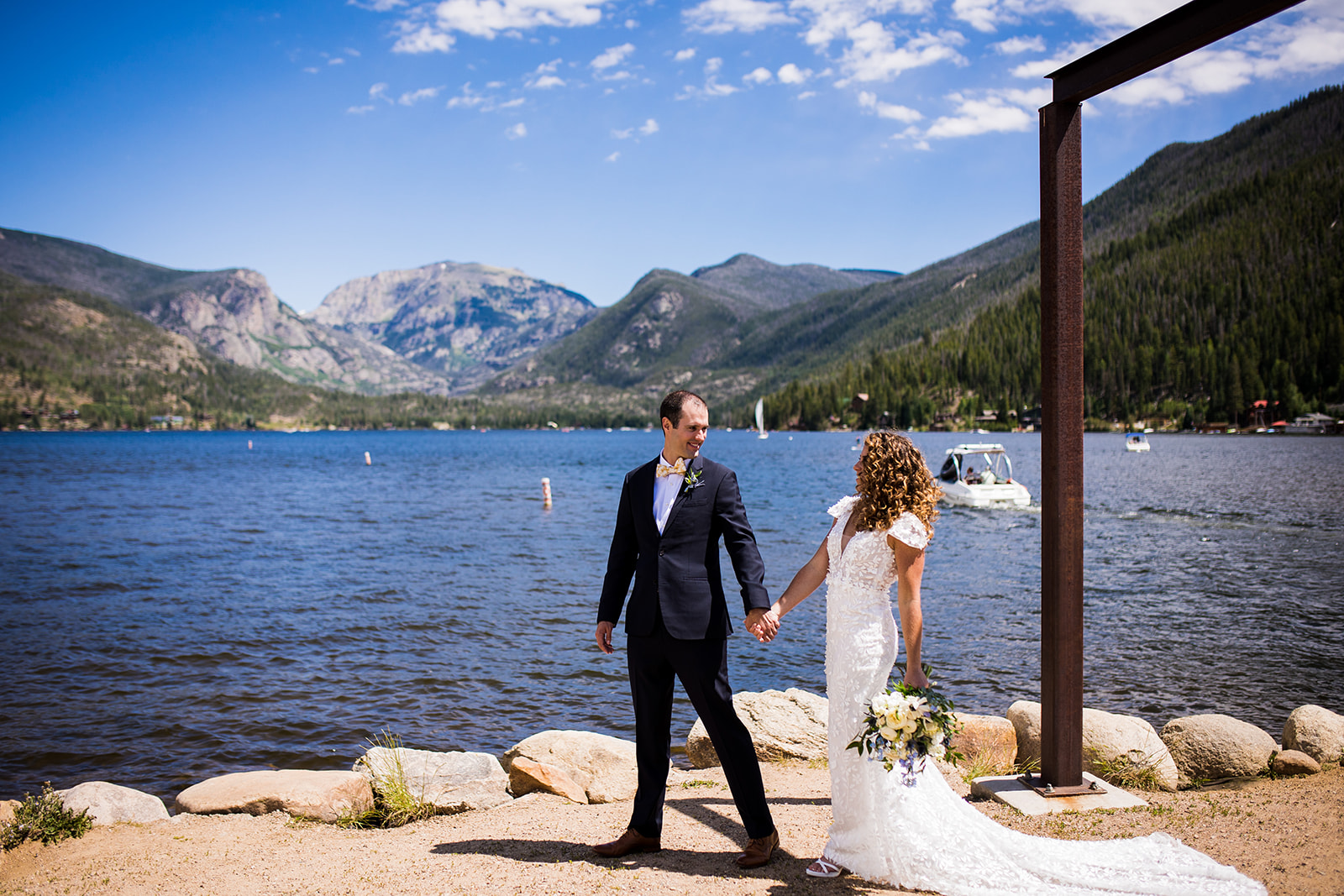 Groom leads bride as the two walk hand in hand with lake and mountain views in the background.