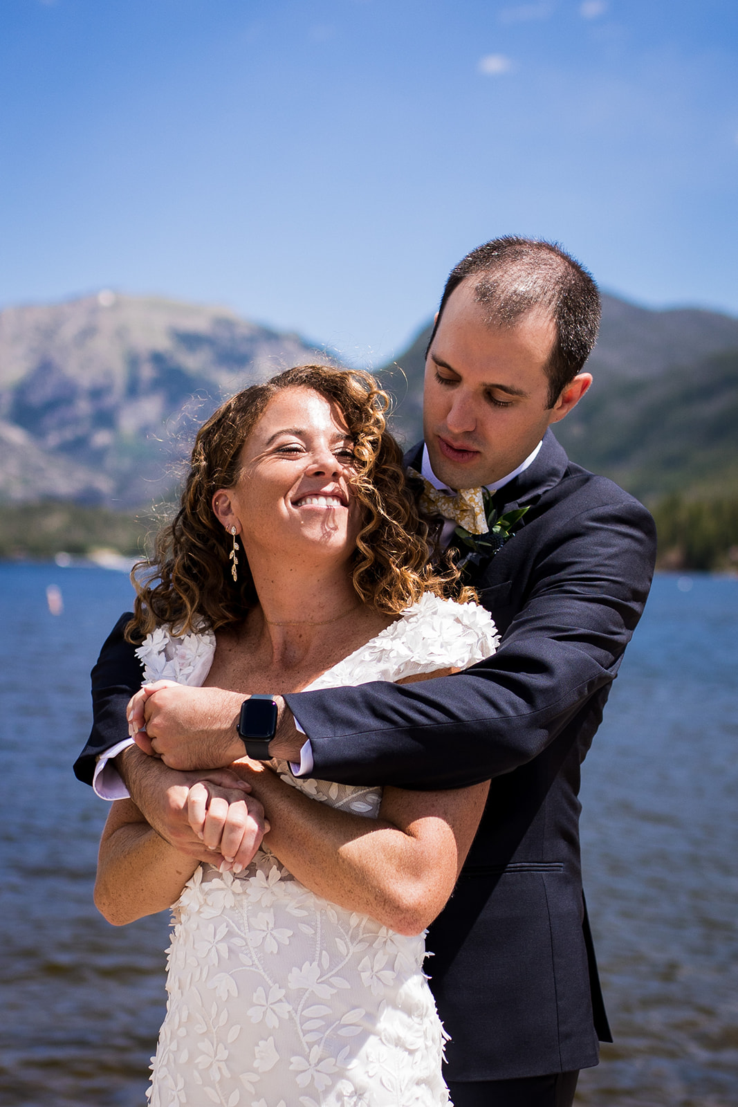 Groom wraps up bride in a hug from behind as the two smile at each other.