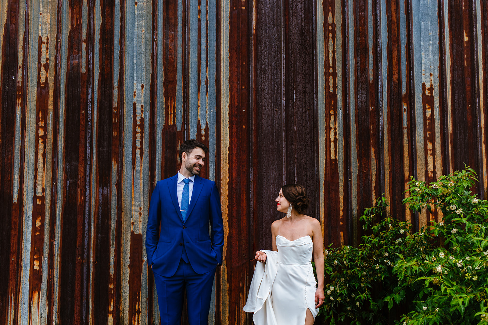 Bride and groom smile and look at each other outdoors against an old factory building.