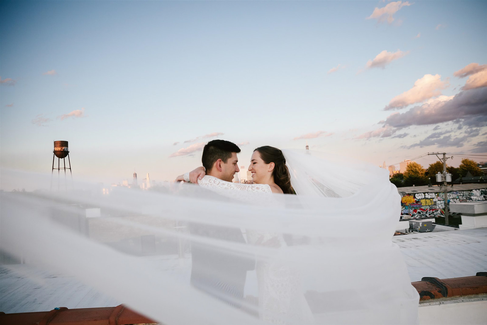 Bride's veil dances in the sunset breeze as she embraces her groom on Walden Chicago rooftop.