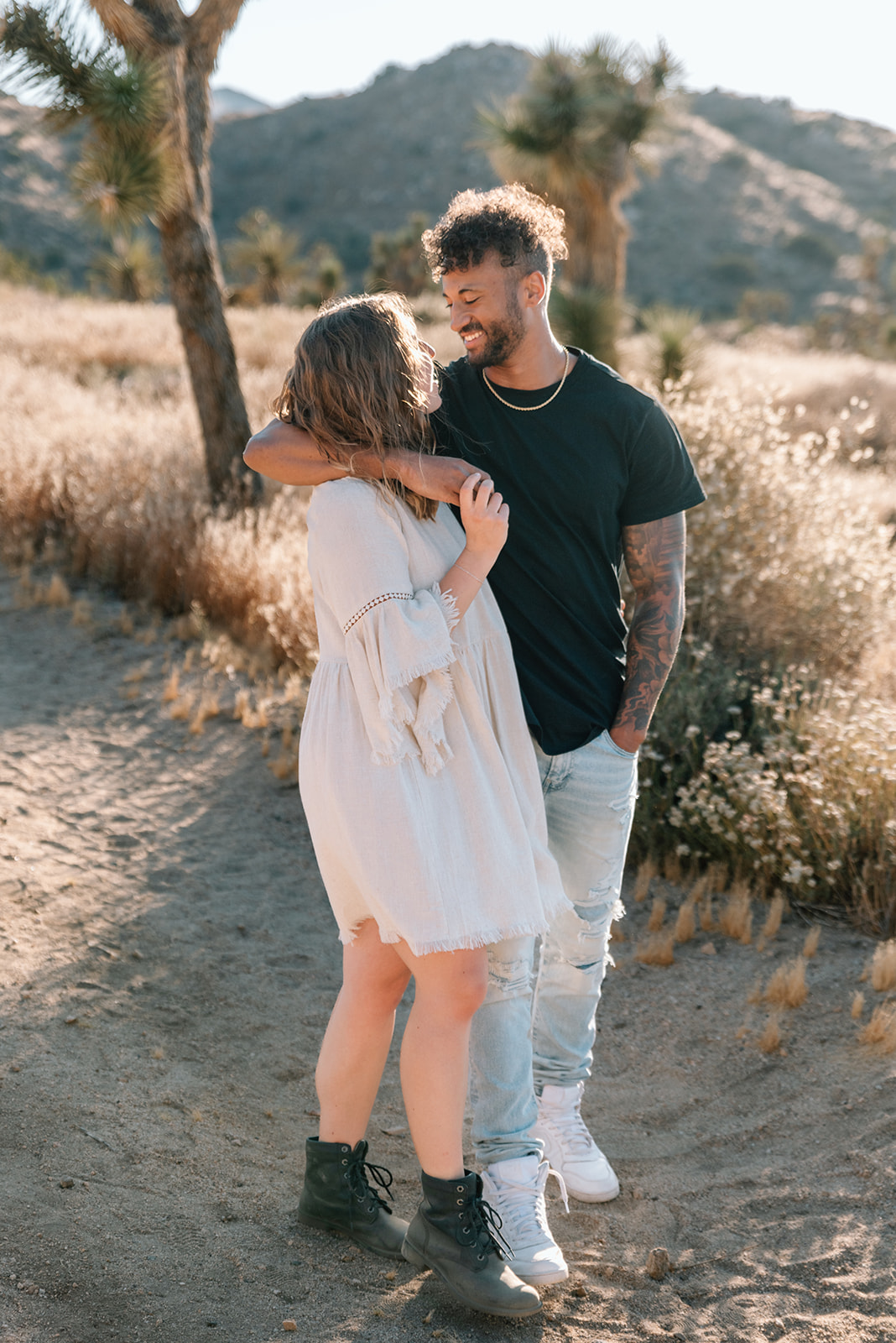 Man casually drapes his arm over his fiance's shoulder as they gaze at each other during their desert engagement session