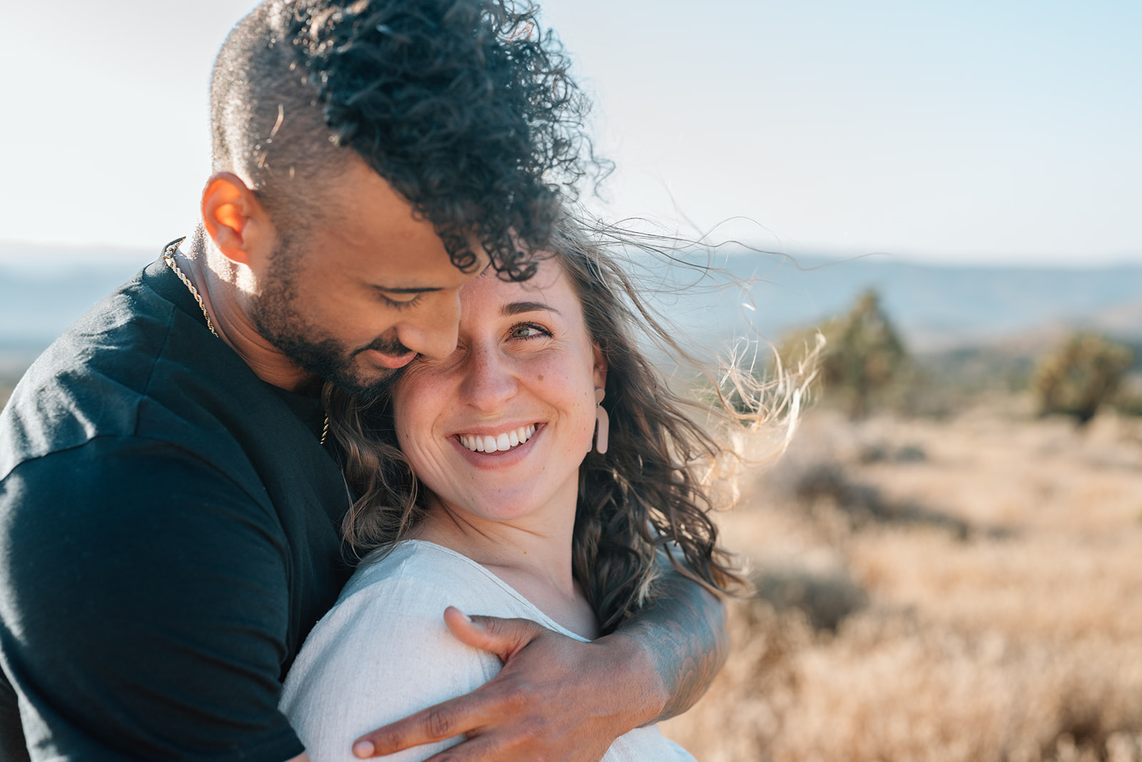 Woman smiles up at man as he hugs her from behind during their desert engagement session
