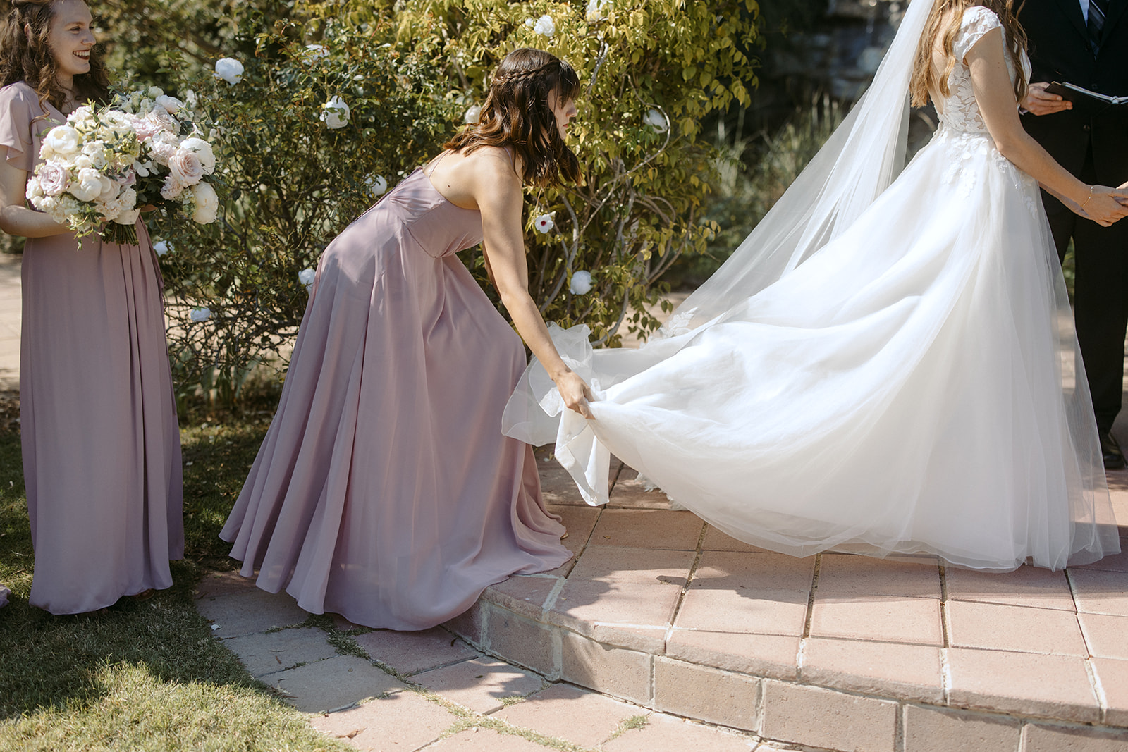 Bridesmaid fluffing bride's dress during wedding ceremony at La Arboleda in California
