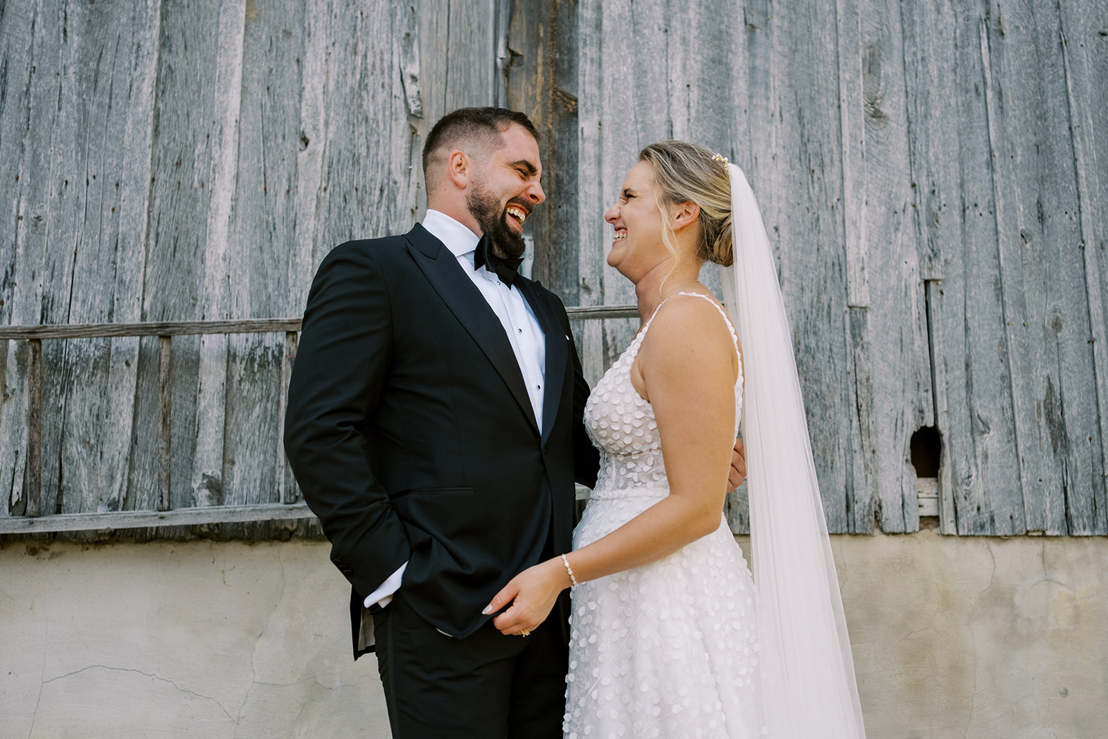 Bride and groom laugh together in front of an old barn at Virginia wedding
