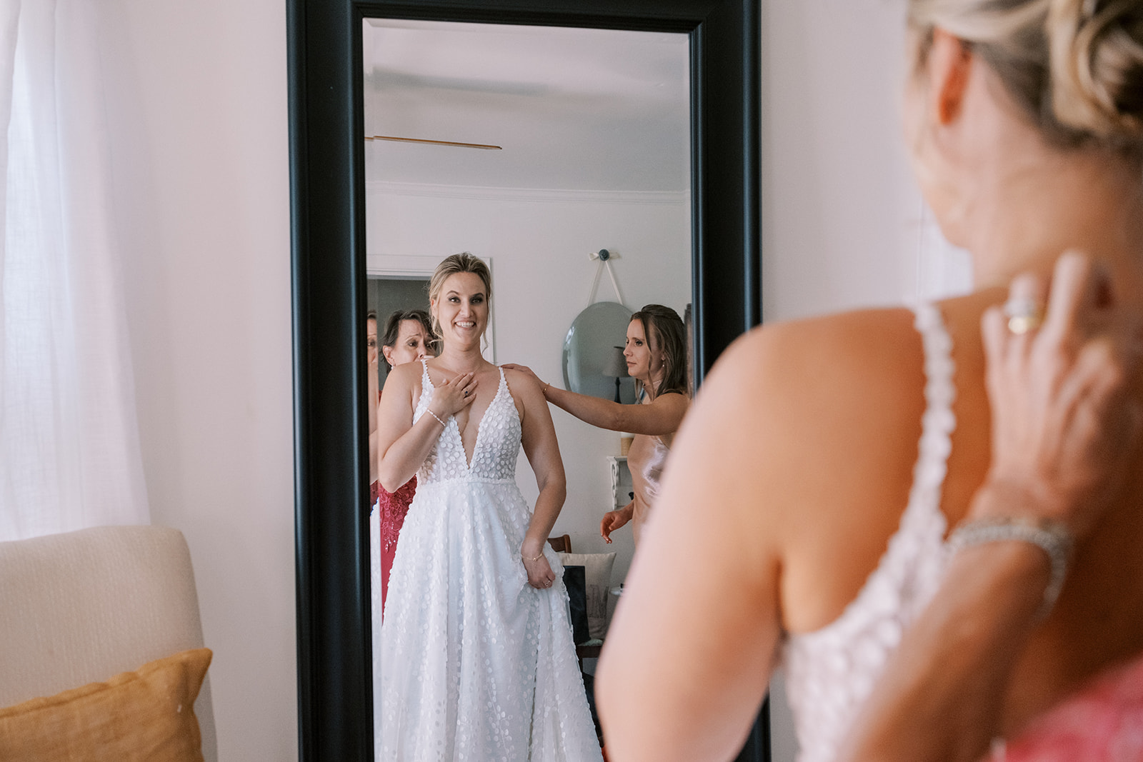 Bride looks at herself in the mirror as bridesmaids button up her dress