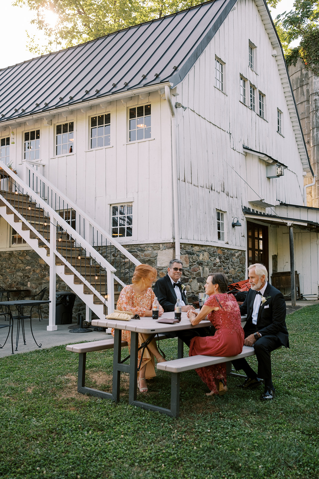 Parents of the bride and groom sit at a picnic table in front of an old barn before the wedding ceremony