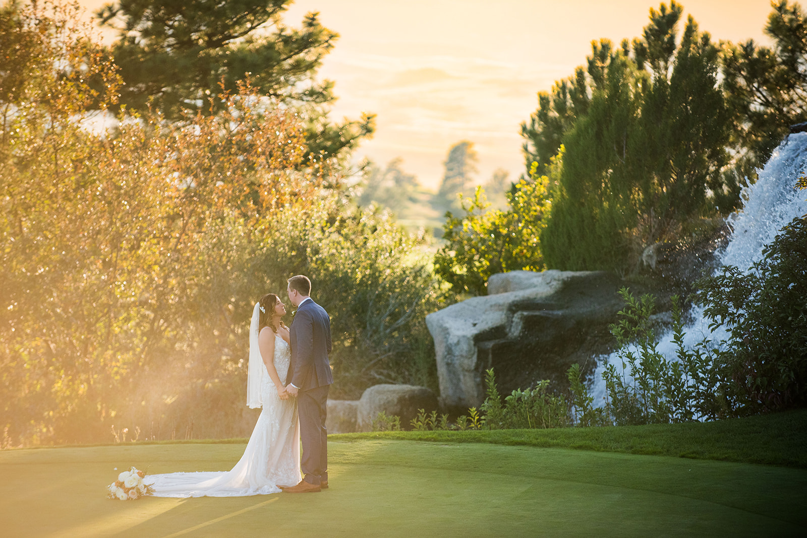 A wide angle shot of the bride and groom holding hands on the gold course at golden hour with the waterfall in the back.