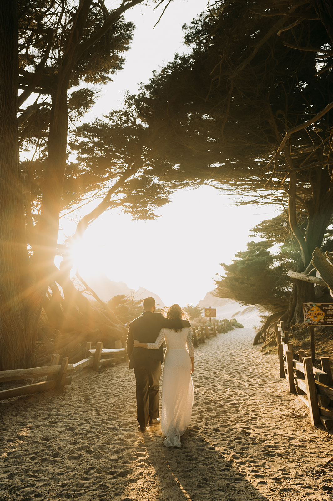 Newly married couple walking on a path to the beach at Big Sur, California with the sun setting over the Pacific Ocean.