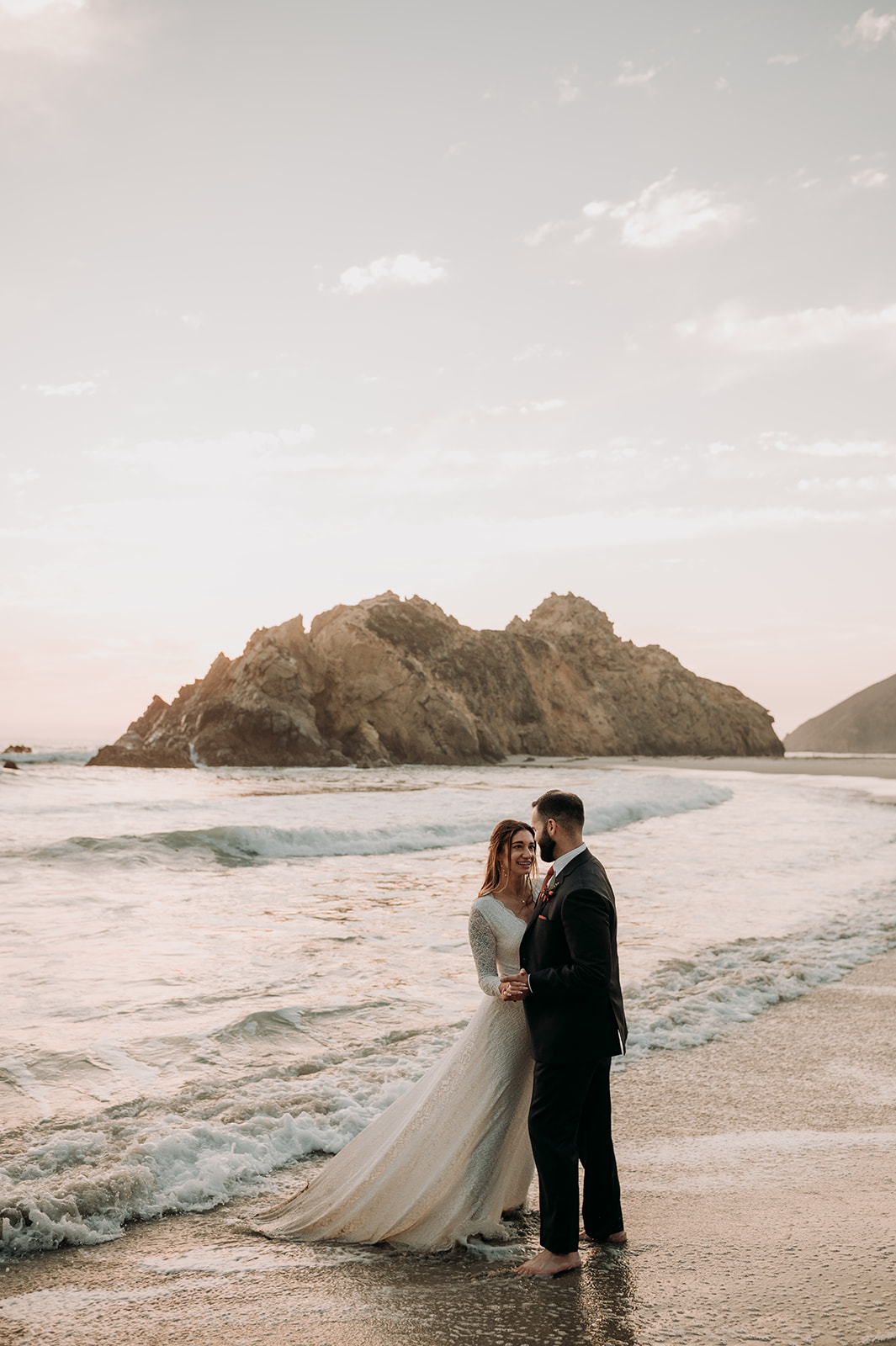Bride and Groom dancing in the waves in wedding attire as the sunsets in Big Sur, California.