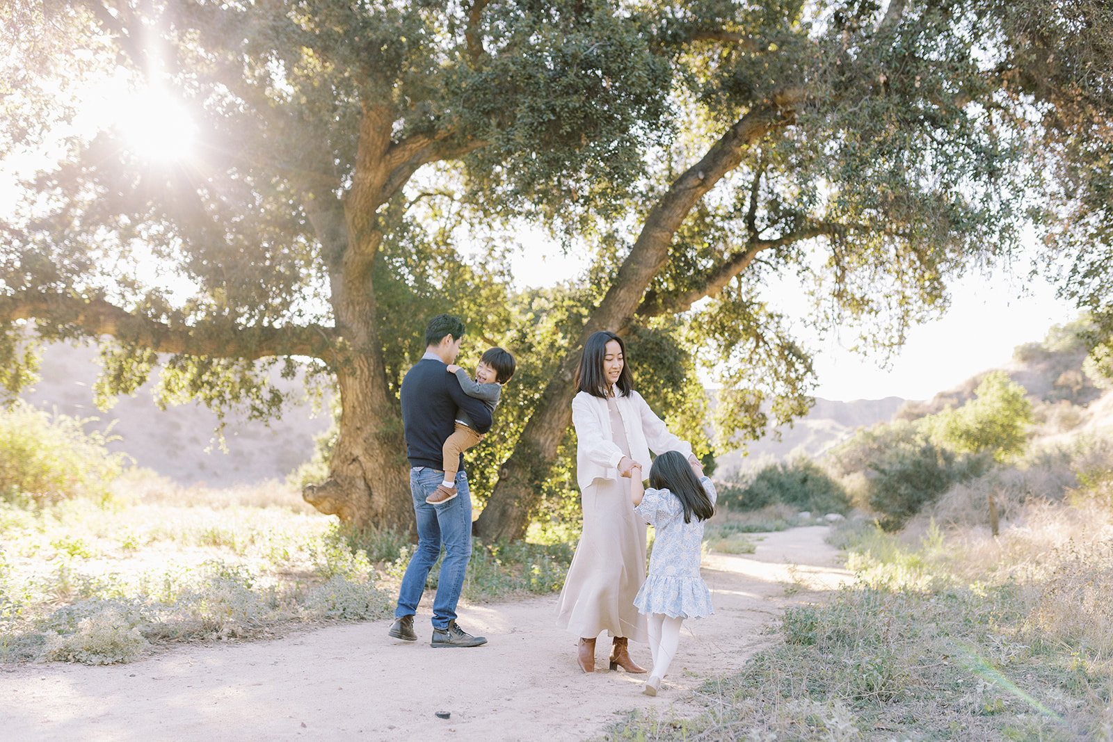 Family with two young children dancing and playing together on a dirt path with a large oak tree behind them.