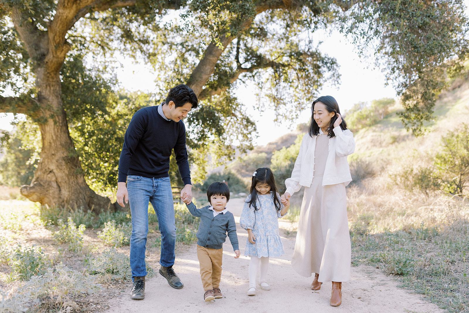 Family with two young children walking down a dirt path with a large oak tree behind them.